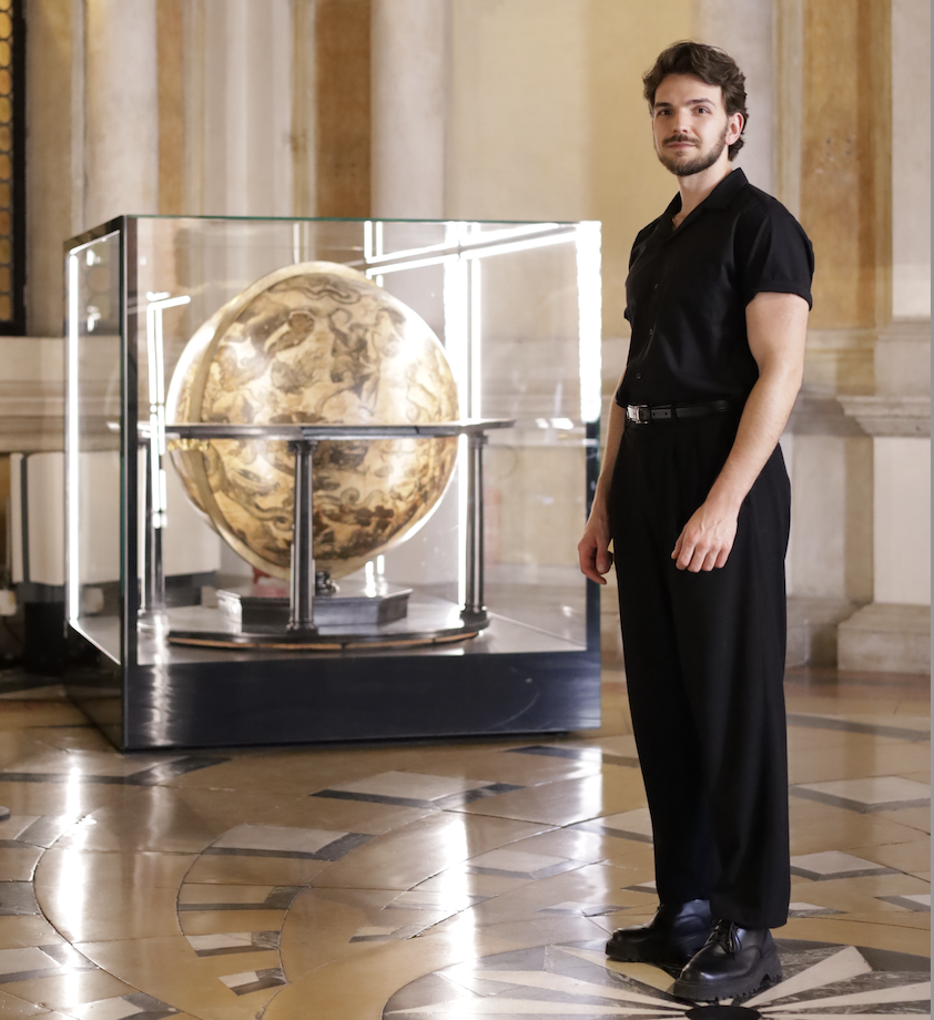 Leonardo Frigo stands in the Marciana Library beside a celestial globe by Vincenzo Coronelli, displayed in a glass case within the ornate marble hall.