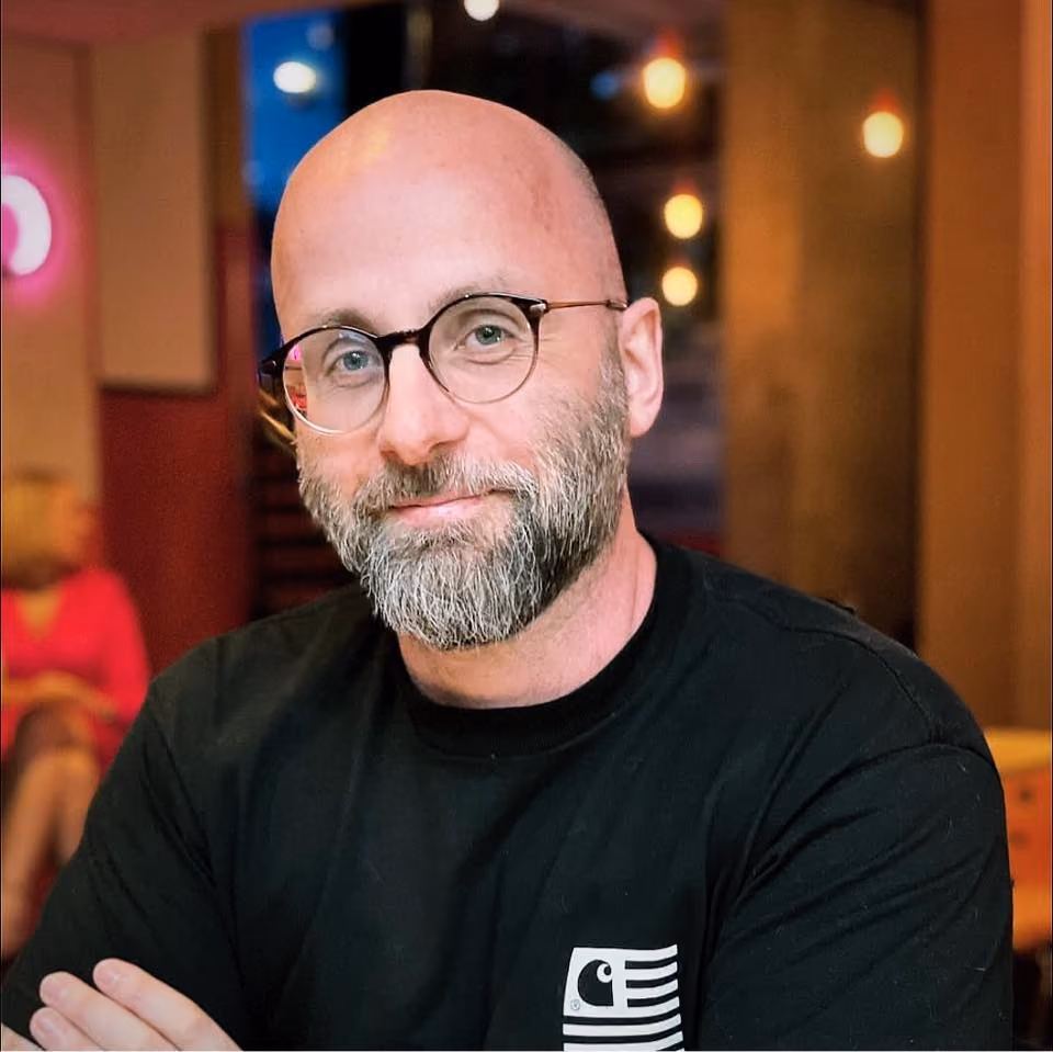 A photo of Jon, wearing a black t shirt with his arms folded. He is smiling and has a full beard and is wearing glasses.