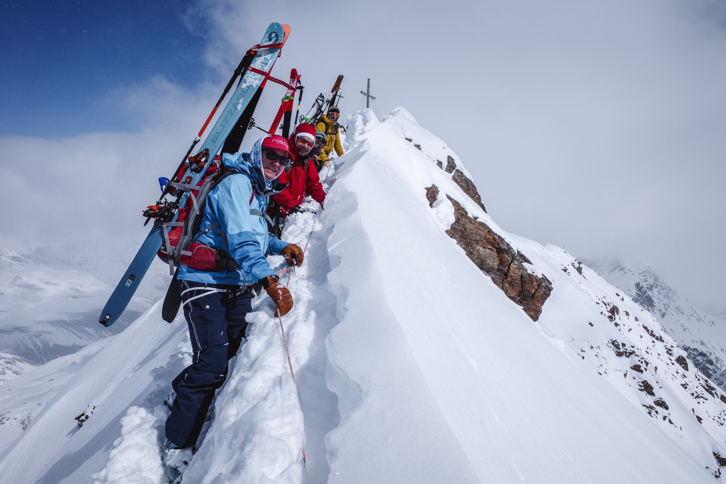 Skiers climbing a snowy alpine ridge toward a summit cross with skis on their packs