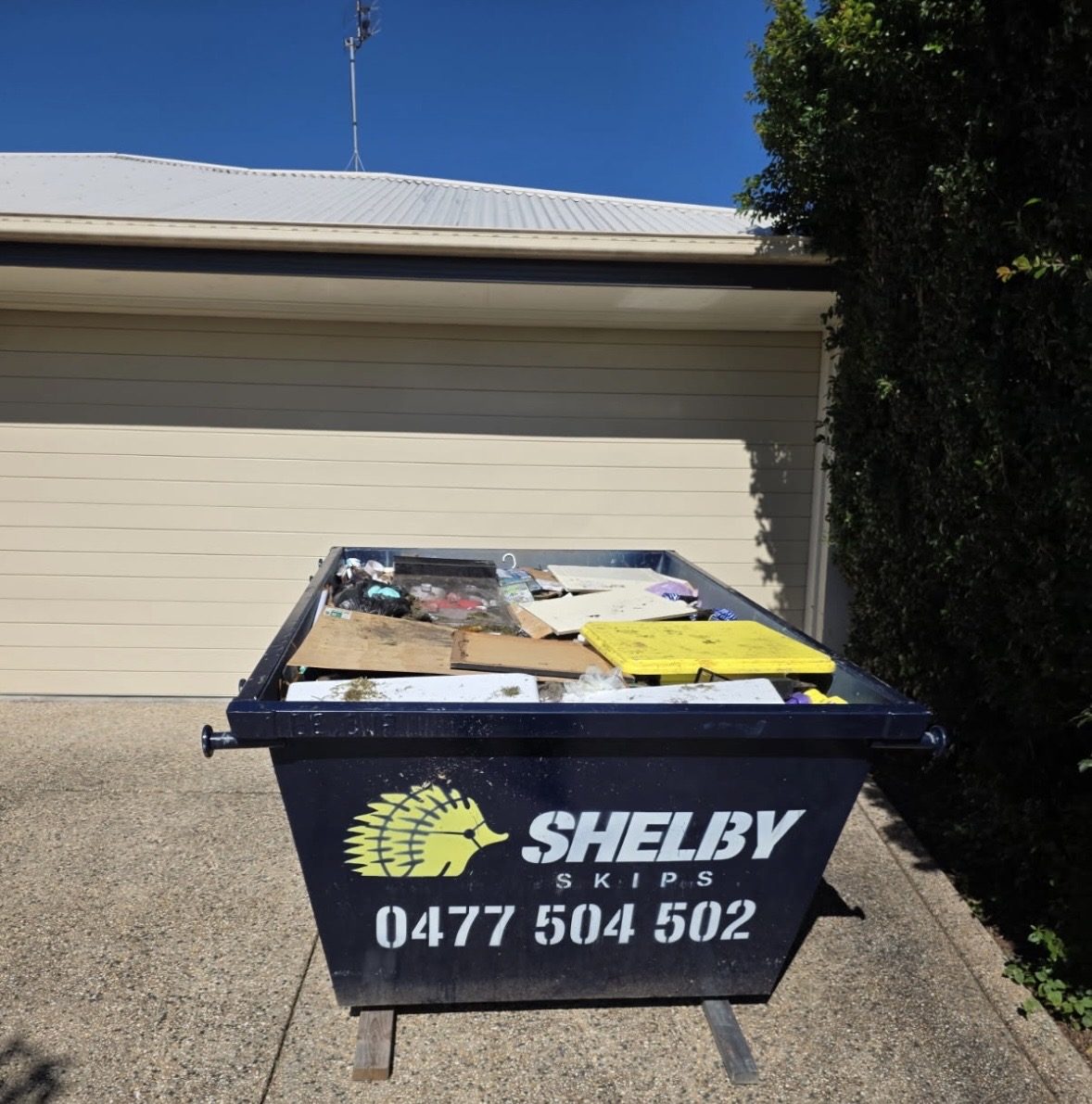 A Shelby Skips bin of household rubbish beside a hedge