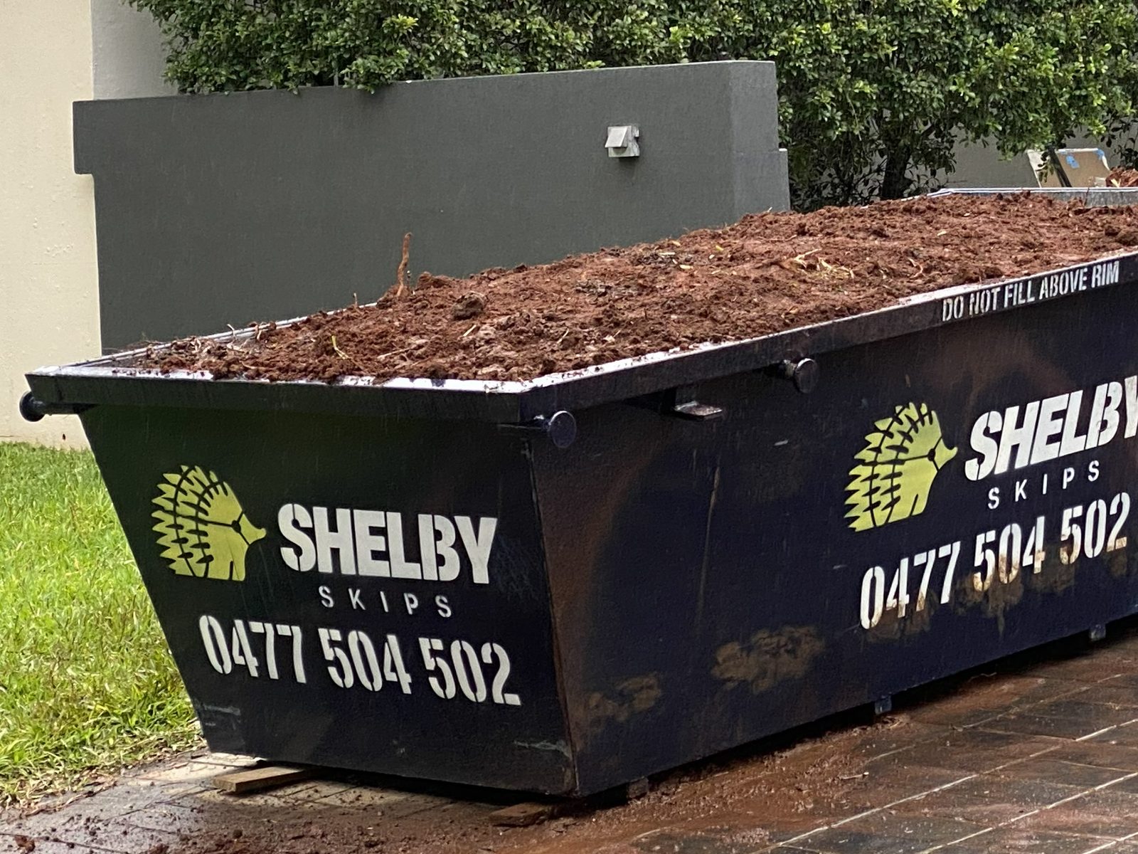A Shelby Skips bin of brown soil on a paved driveway, with a hedge and a garden wall behind.