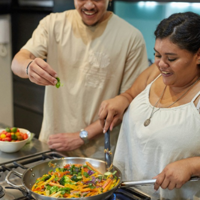 Two people smiling as they cook a vegetable stir-fry together in a pan on a stovetop.