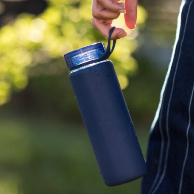 A hand holding a navy reusable drink bottle outdoors, with sunlit greenery blurred behind.