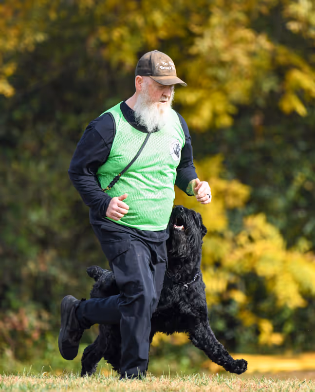 Bearded man wearing a green vest and cap jogging with a black dog on grass with trees in the background.