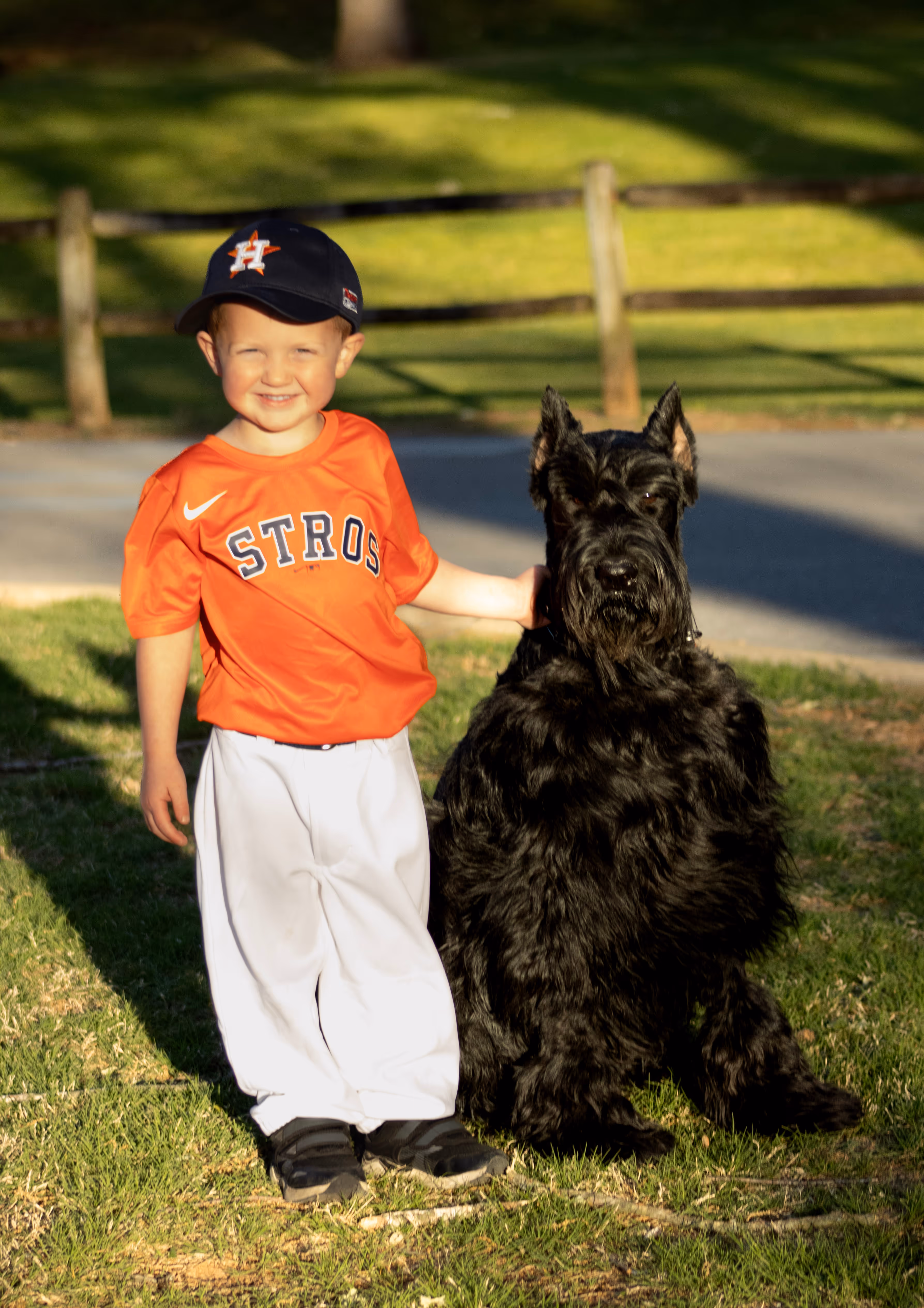 Smiling young boy in an orange Astros baseball jersey and white pants stands on grass with one arm around a large black dog.