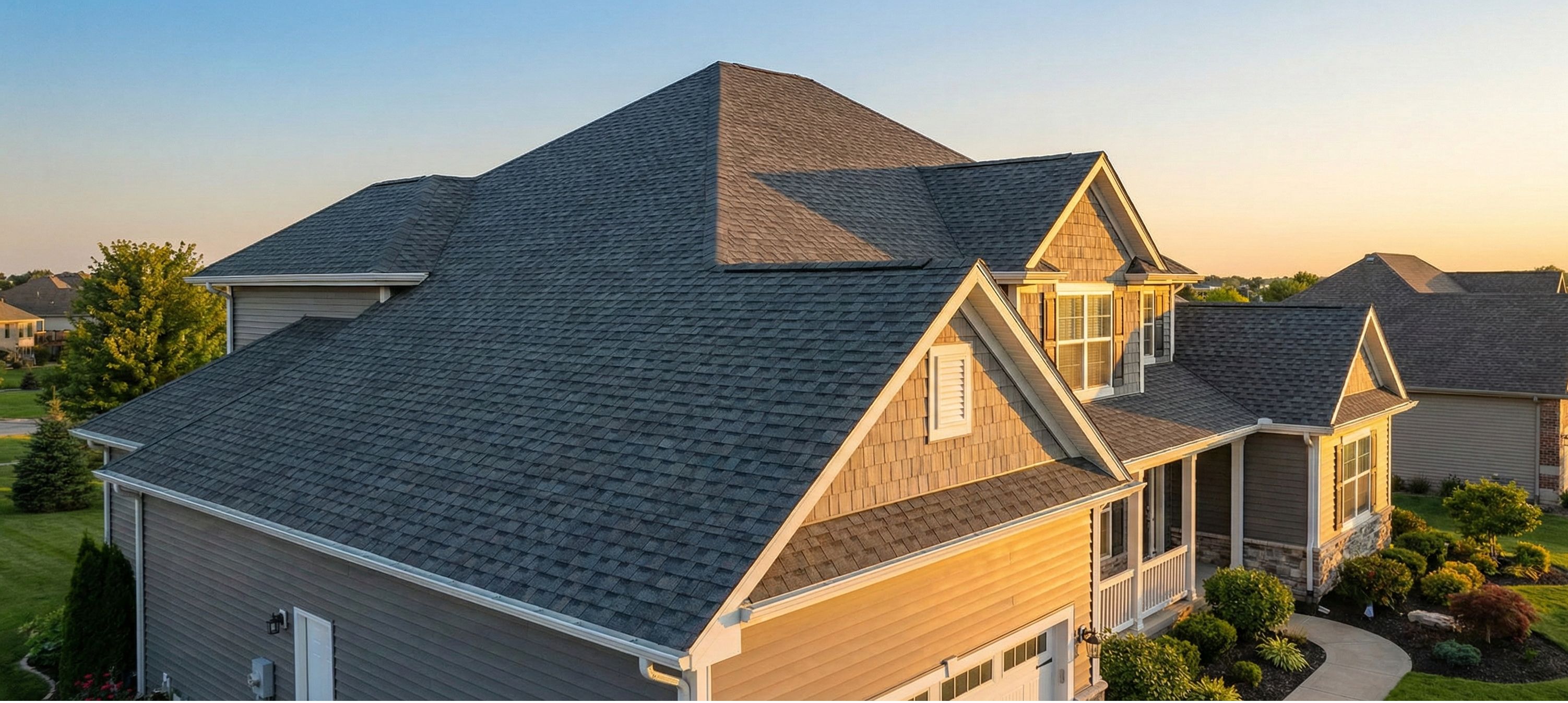 Modern two-story house with gray shingle roof, beige siding, and manicured lawn at sunset.