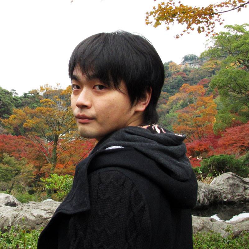 Ryo Kanda with black hair wearing a black jacket stands outdoors in front of colorful autumn trees and rocks.