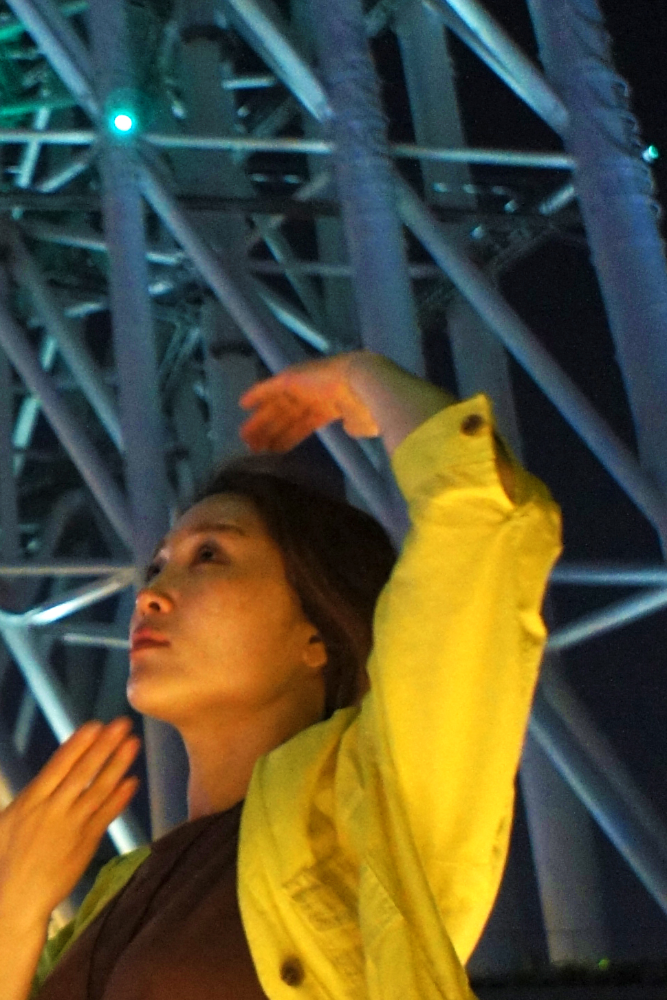Aiko Hatanaka in a yellow jacket poses with one arm raised in front of a large metal structure at night.