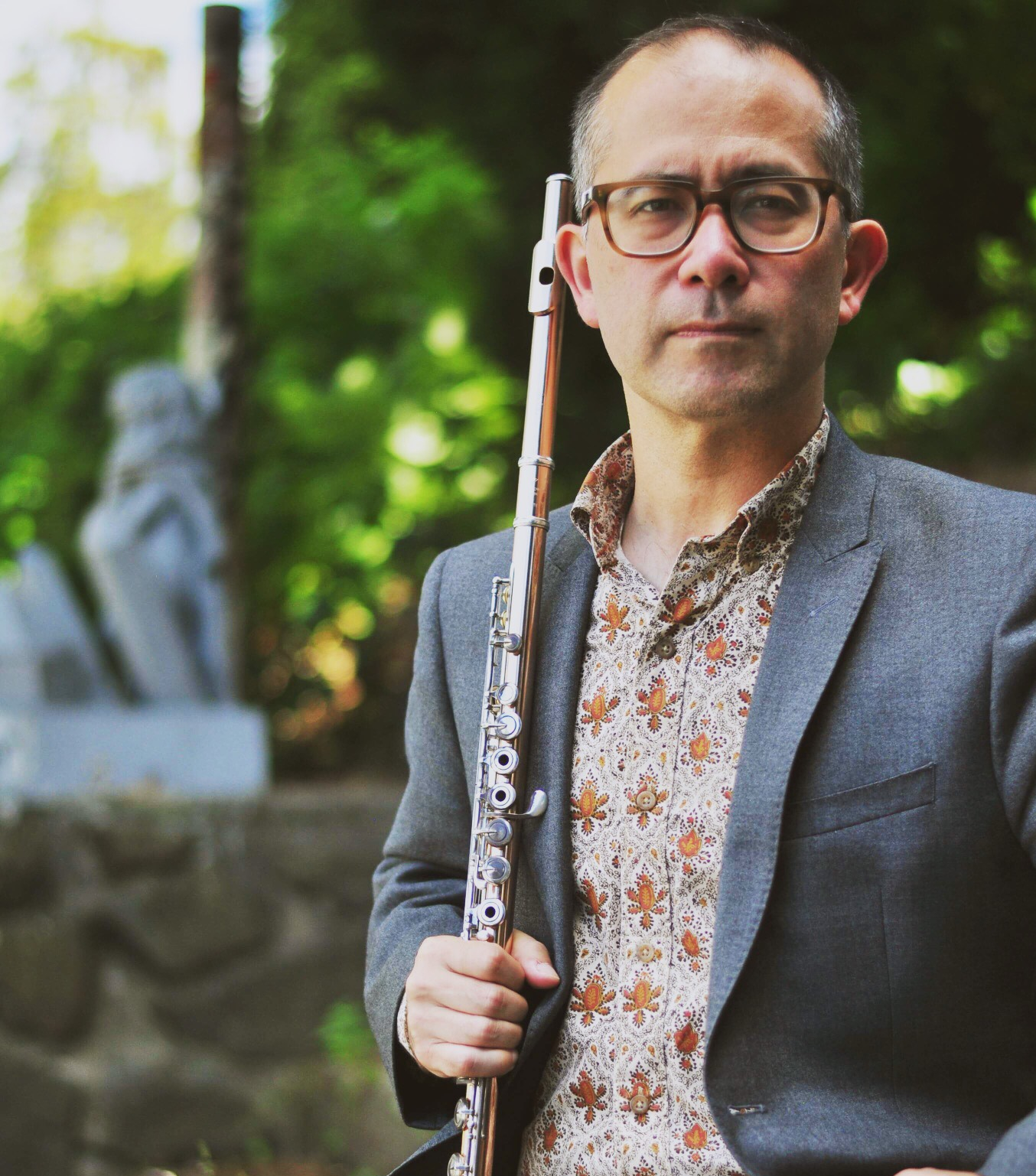 Mark McGregor wearing glasses and a patterned shirt under a gray blazer holds a flute outdoors, with greenery and a stone sculpture in the background.