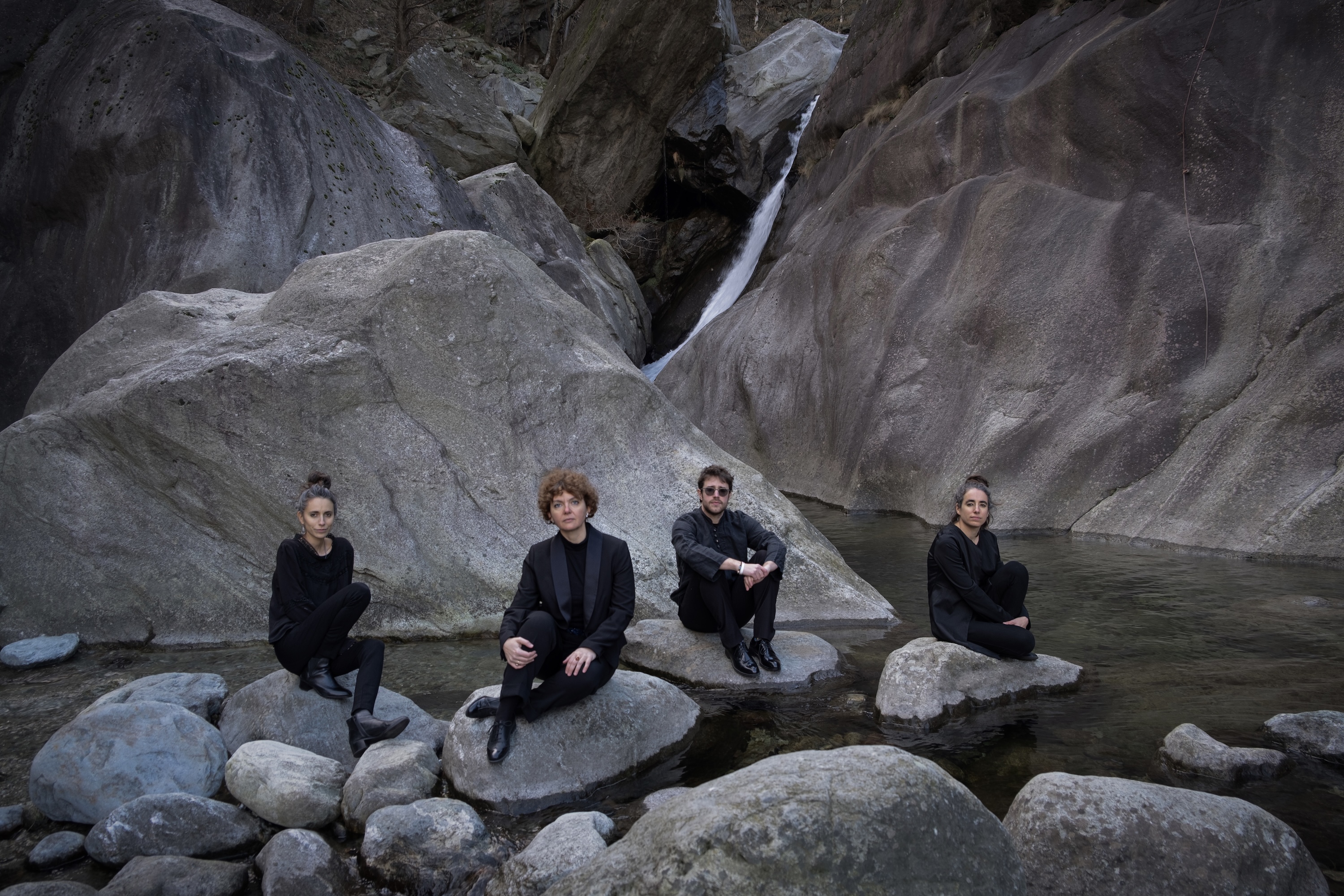 Quartetto Maurice's members in black clothing sit on rocks by a stream with large boulders and a small waterfall in the background.
