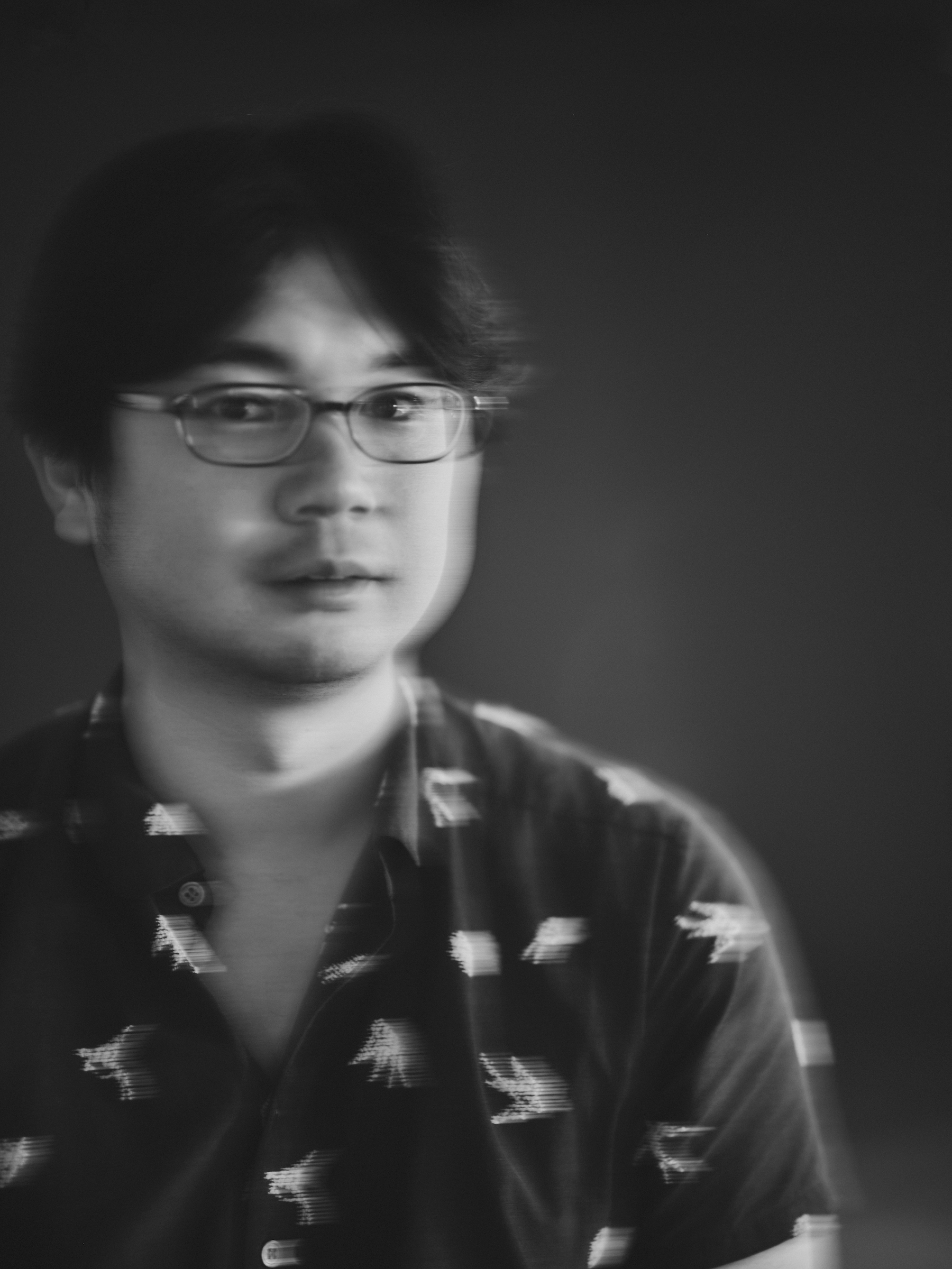 Rurihiko Hara with glasses and dark hair wears a patterned shirt and looks toward the camera; the photo is in black and white with a blurred, double-exposure effect.