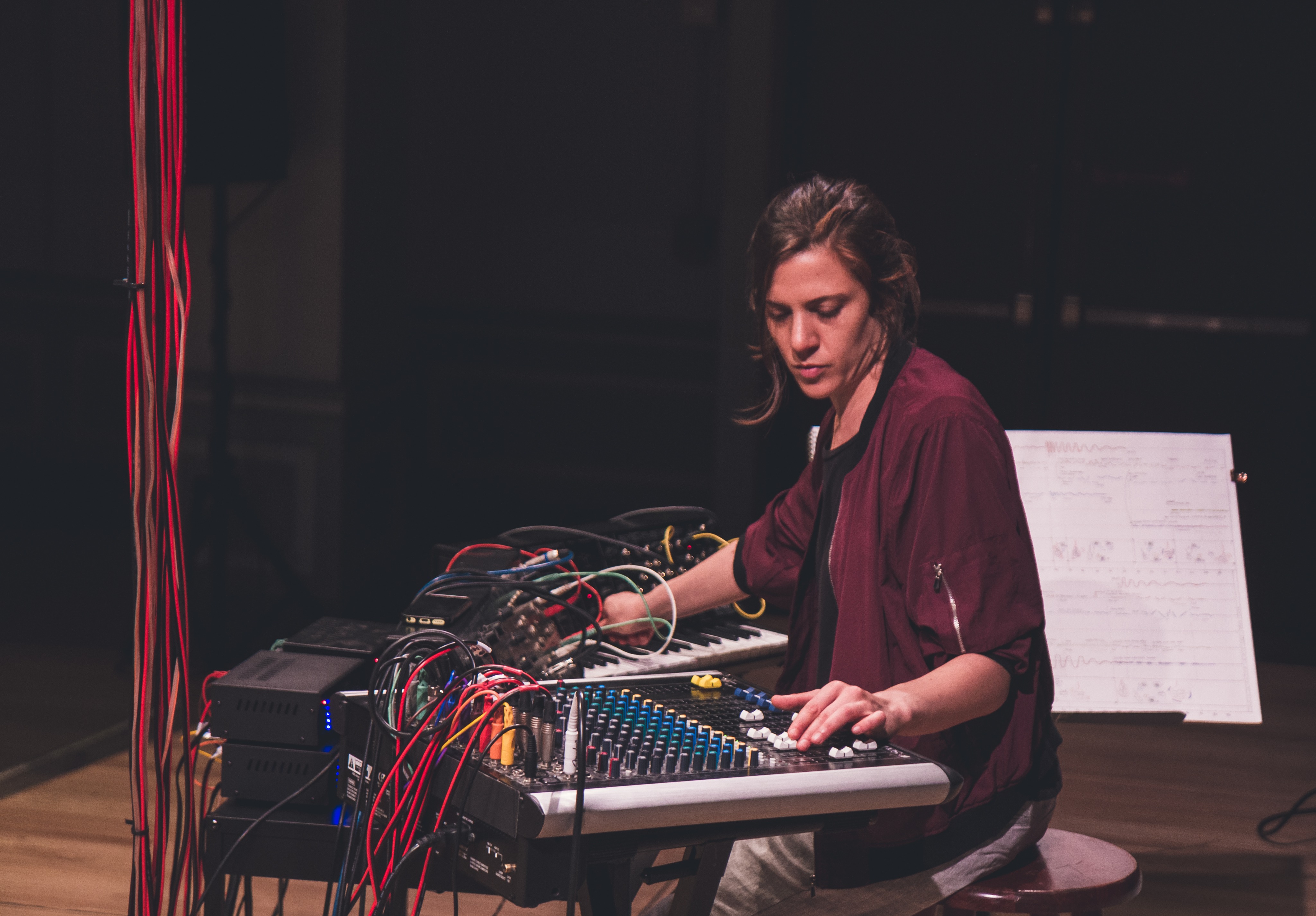 Cecilia Lopez sits sideways on the right playing a keyboard and synthesizer with a column of red cables to the left.