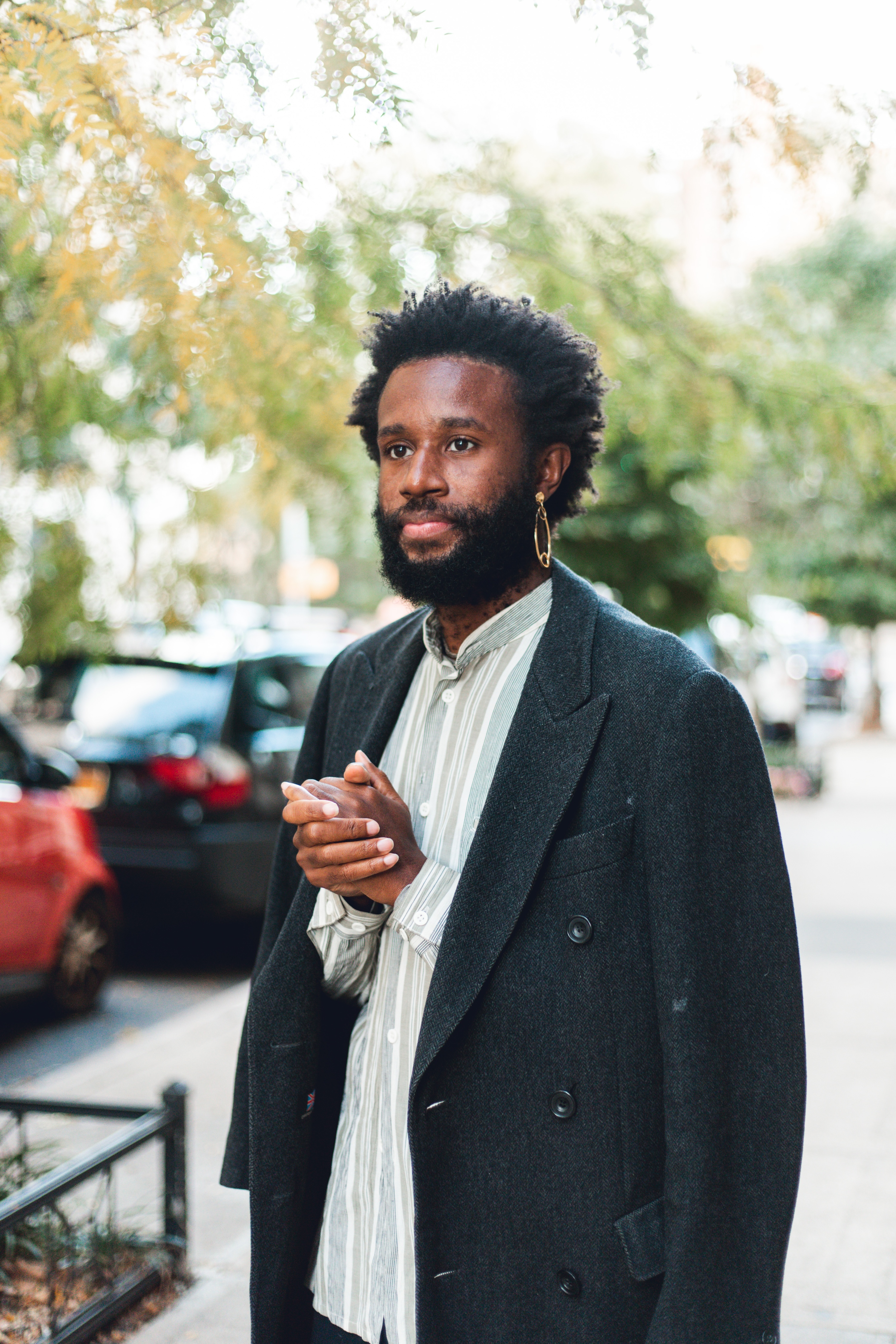 Jerome Ellis stands at centre, turned slightly to his right, with both hands clasped at his chest in a striped button-up shirt and dark blazer draped over his shoulders