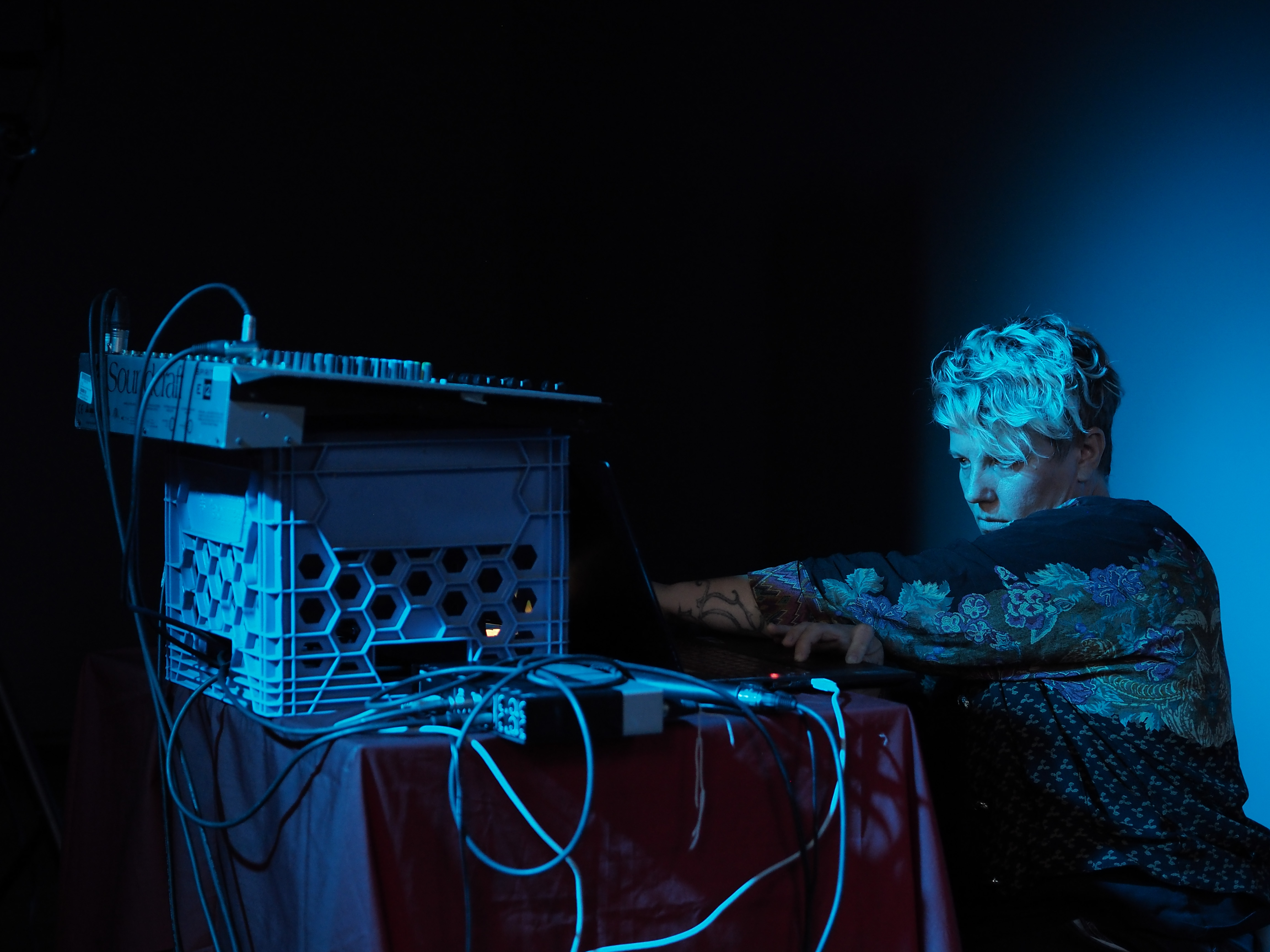 AO Roberts with short, curly hair operates electronic music equipment on a table under blue lighting.