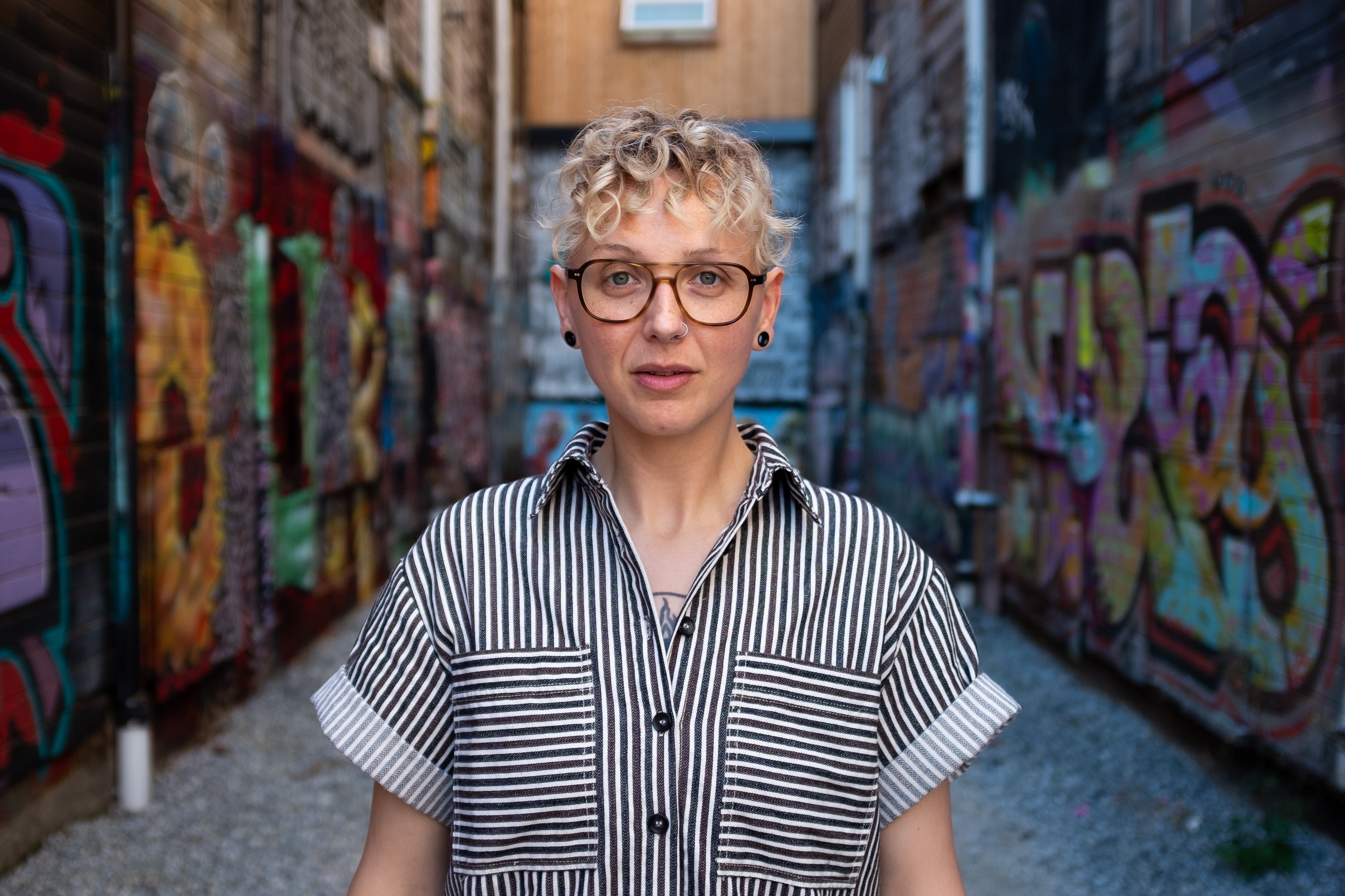 SJ Kirsch with short curly hair, glasses, and a striped shirt stands in an alleyway with colorful graffiti on the walls.