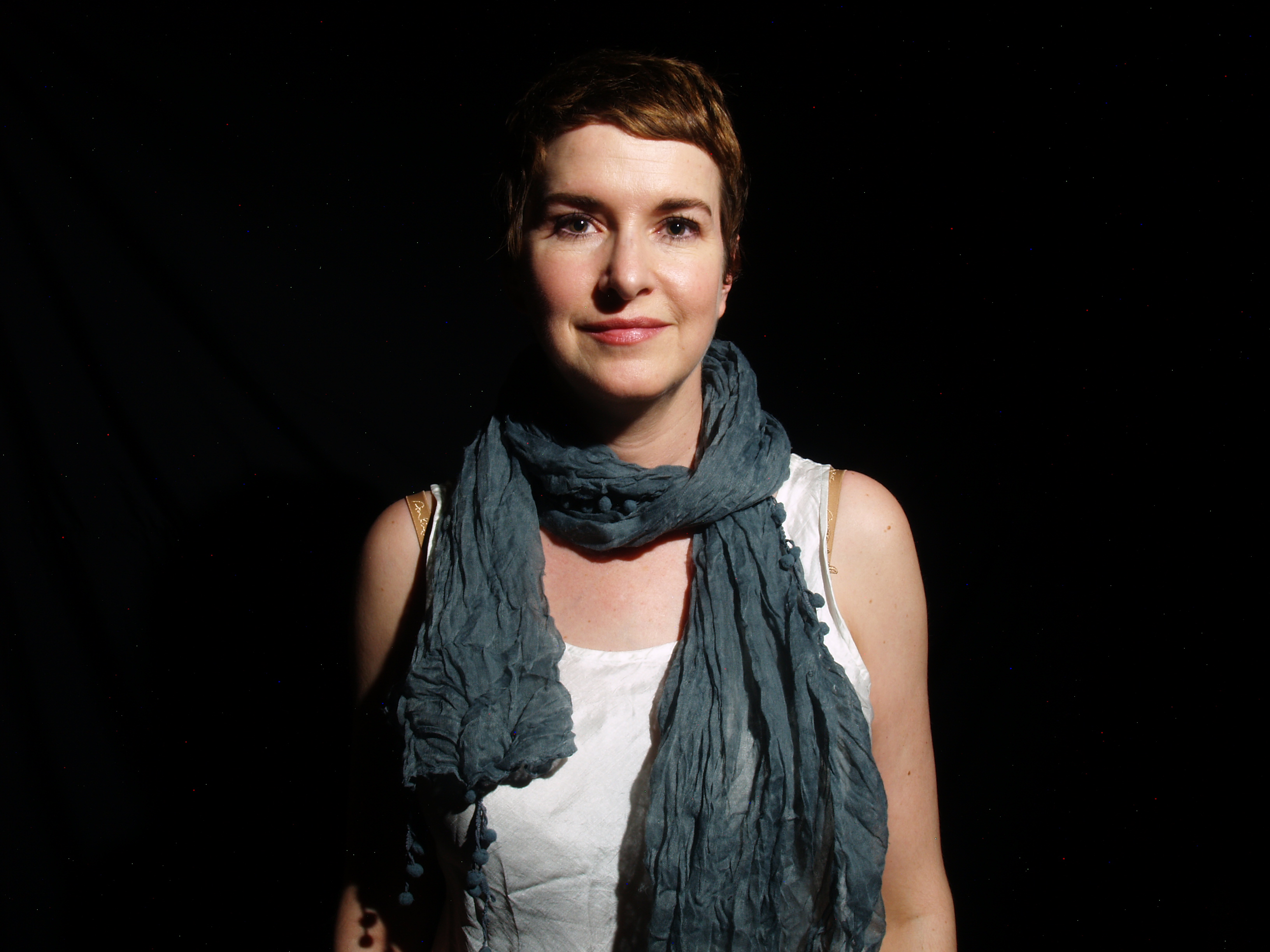 Viviane Houle with short brown hair wearing a white top and a gray scarf stands against a black background, looking at the camera.