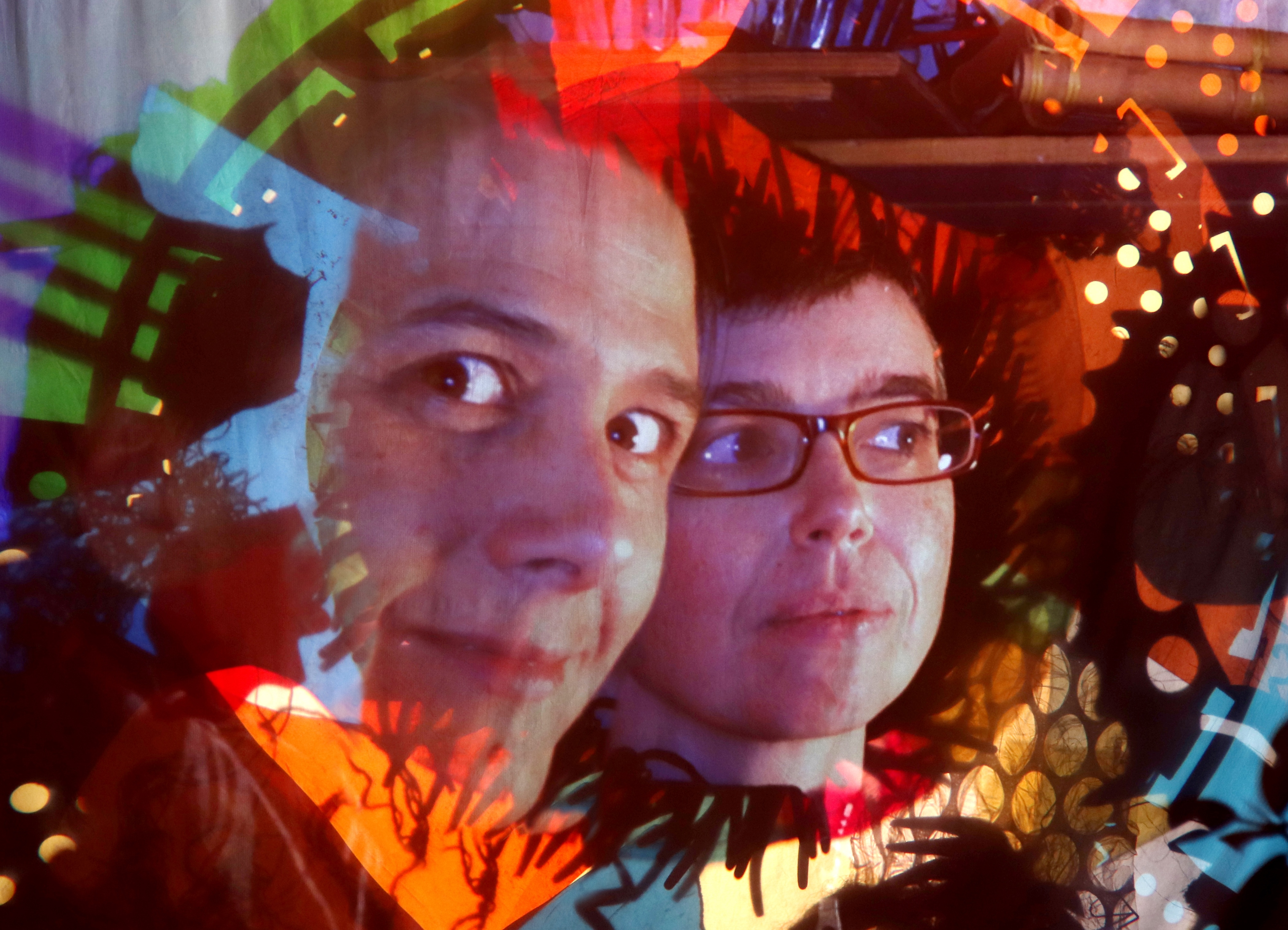 Kaleidescopic fragments of light and colour frame a close-up image of Jessica Gabriel, on the left, looking wide-eyed at the camera, and Chloé Ziner, on the right, wearing glasses and looking askance to their left.