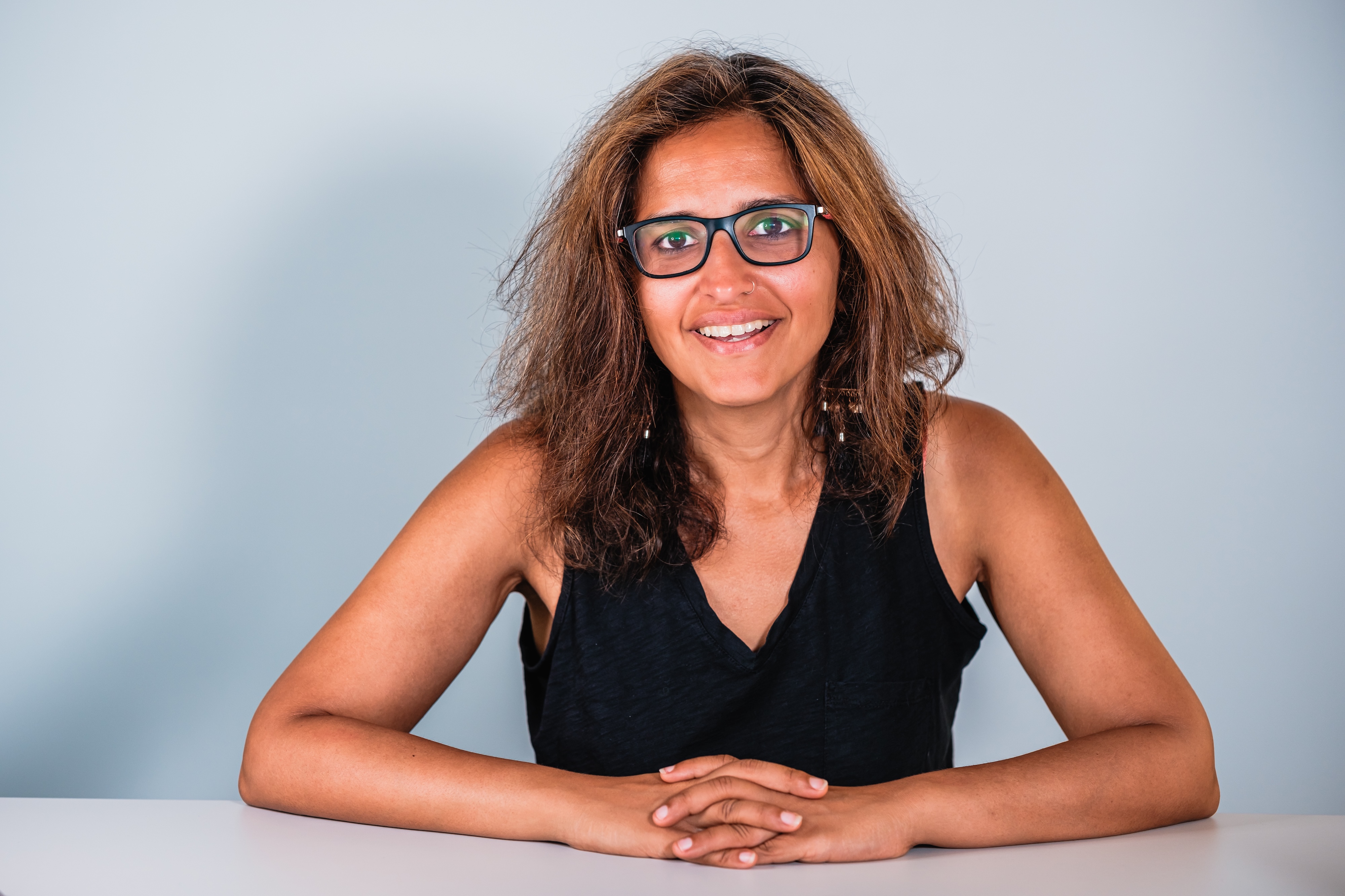  In front of a soft hazy blue background, Harsha Walia, smiling, faces the camera, with her elbows outstretched and hands folded in front of her on top a white surface.
