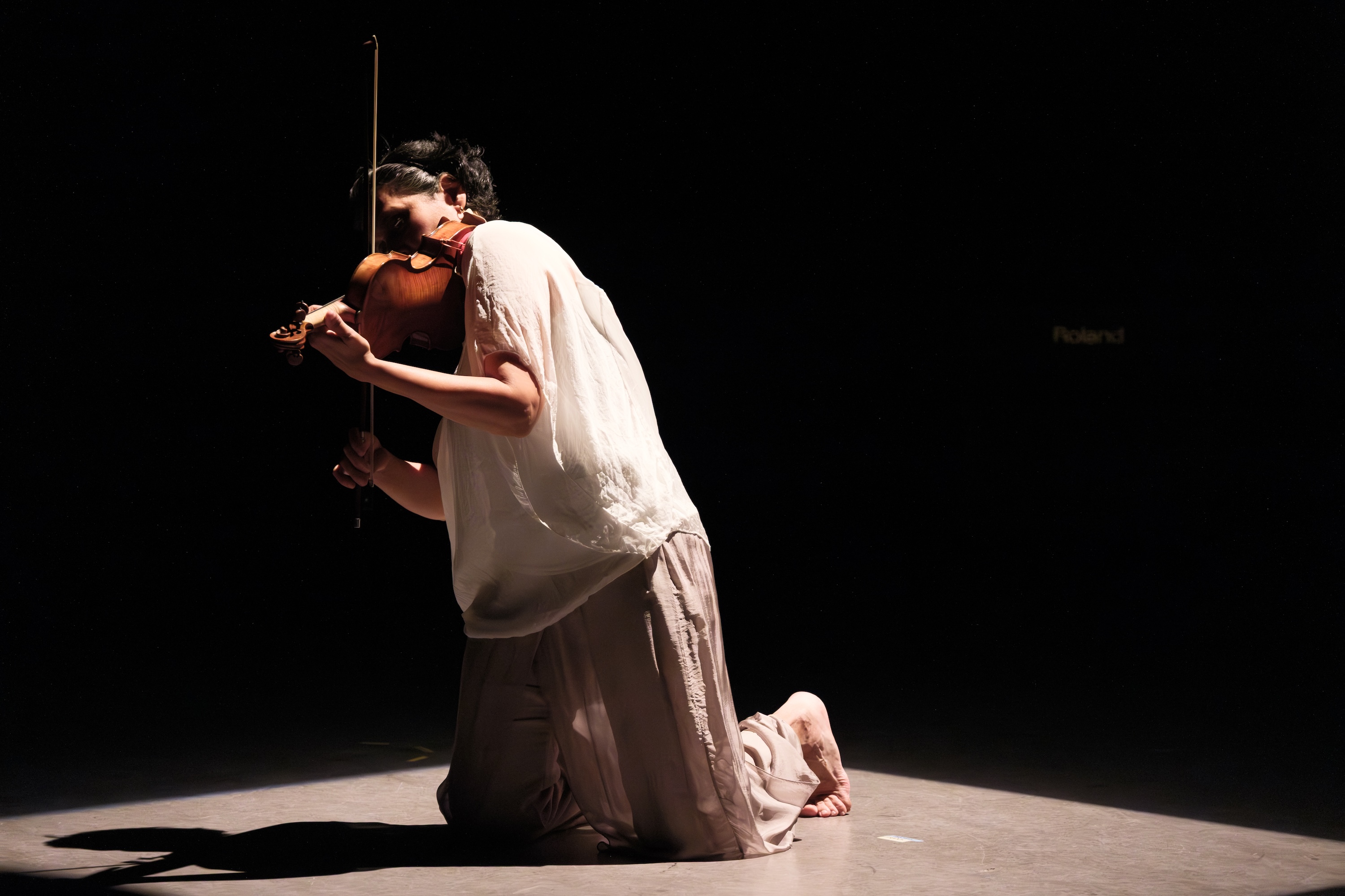 Parmela Attariwala kneels on stage under a spotlight, playing a violin, wearing loose light-colored clothing, with a dark background.