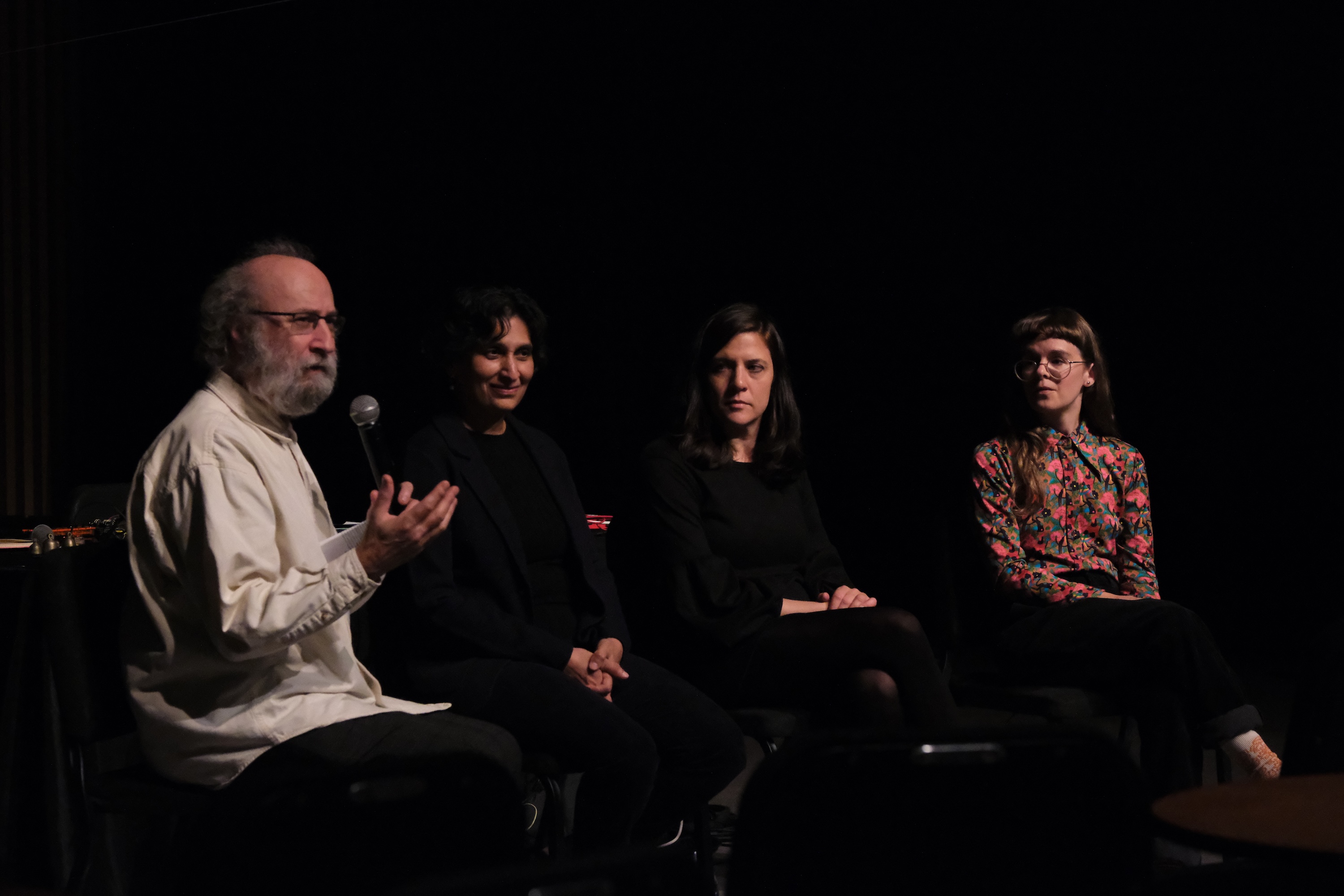 Émilie Payeur, Parmela Attariwala, Cecilia Lopez, and Giorgio Magnanensi are seated on a dark stage; Giorgio on the left speaks into a microphone while Émilie, Parmela, and Cecilia next to him listen attentively.