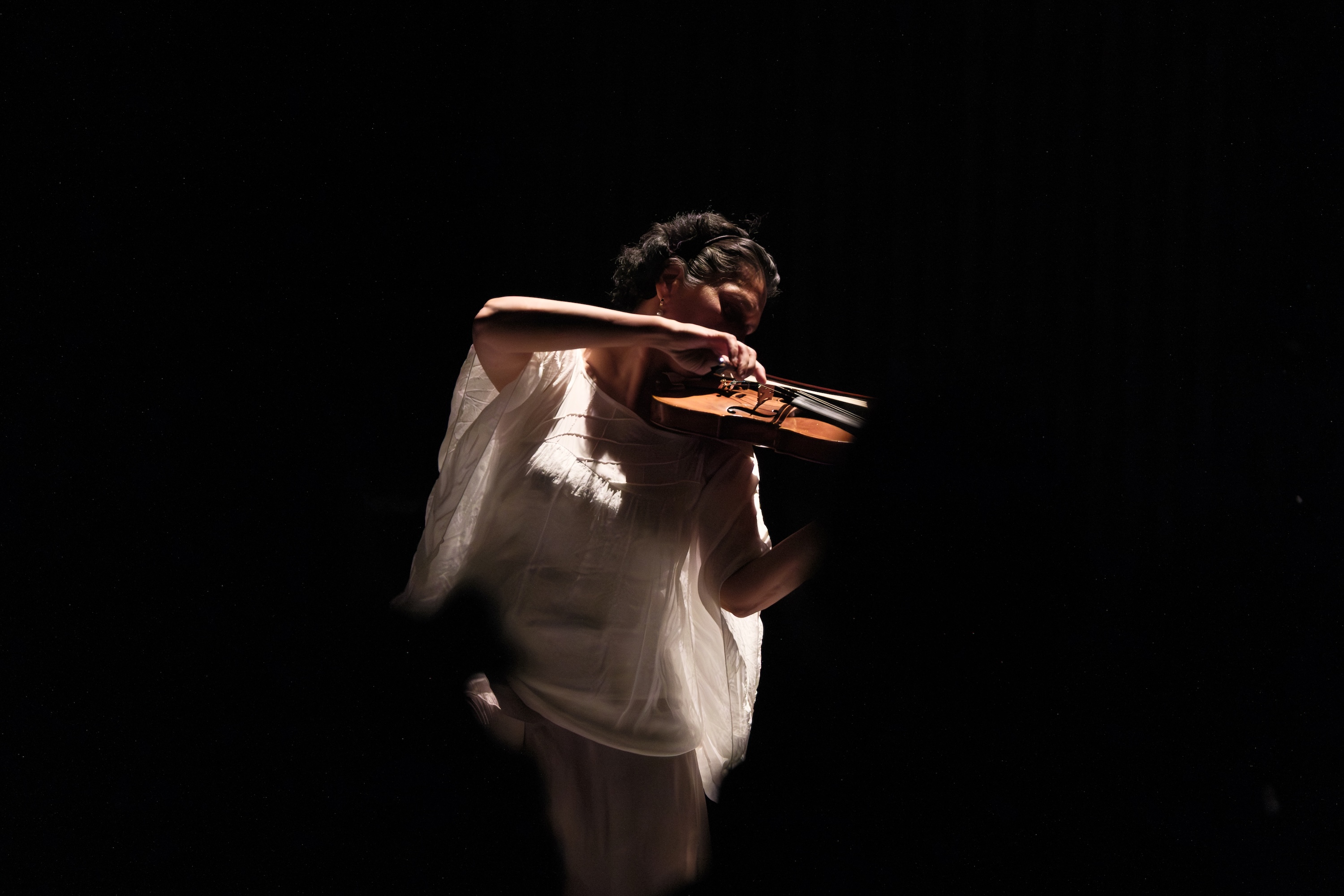 Parmela Attariwala in a white outfit plays the violin on a dark stage, illuminated by a spotlight from above.