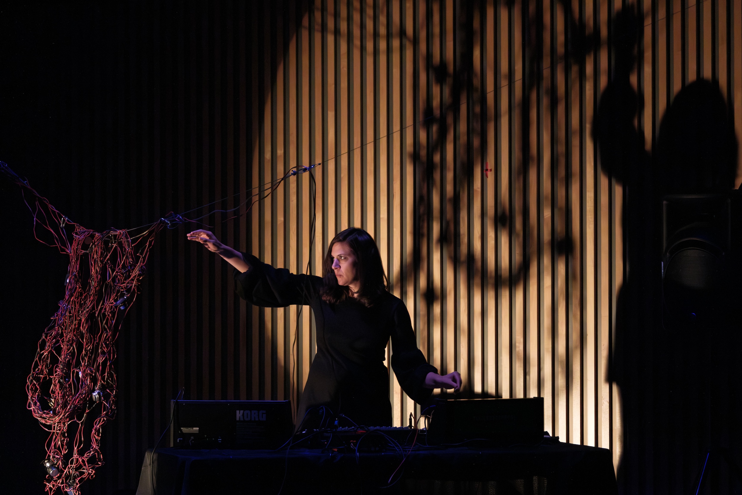 Cecilia Lopez in black operates electronic music equipment on a stage, reaching toward a bundle of cables with a dramatic shadow cast on a wooden wall behind them.