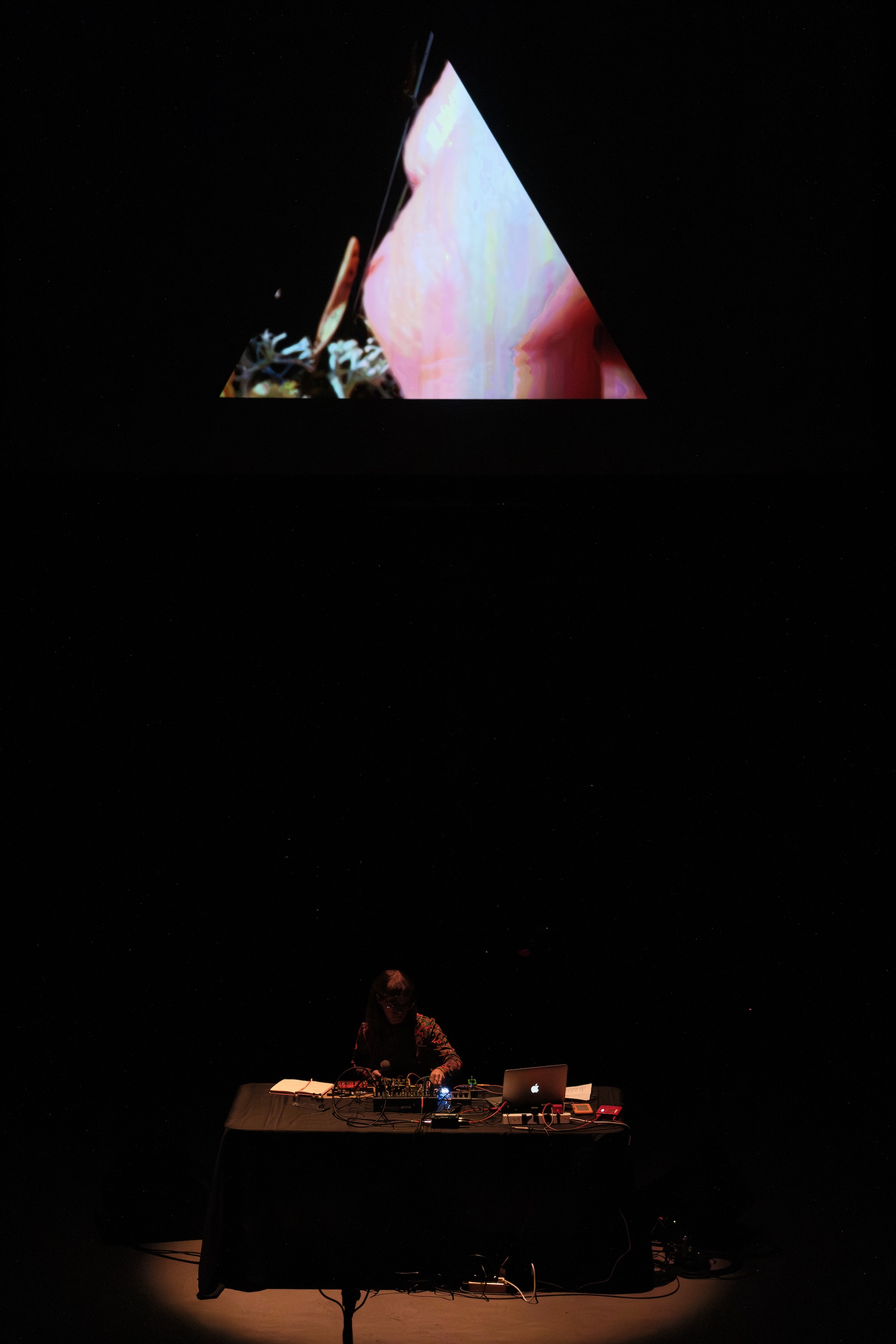 Émilie Payeur sits at a table with electronic equipment onstage, while a triangular video projection is displayed above them in a dark setting.