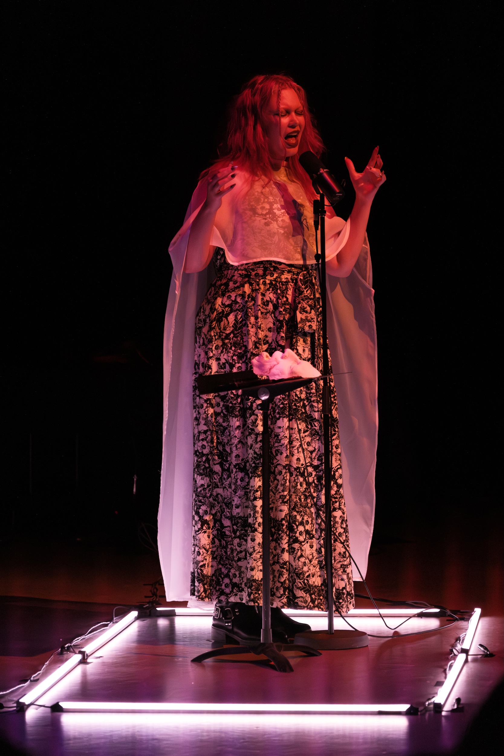 Erin Gee in a floral dress and sheer cape passionately sings into a microphone on stage, with dramatic lighting and a stand holding a white mask in front of them.