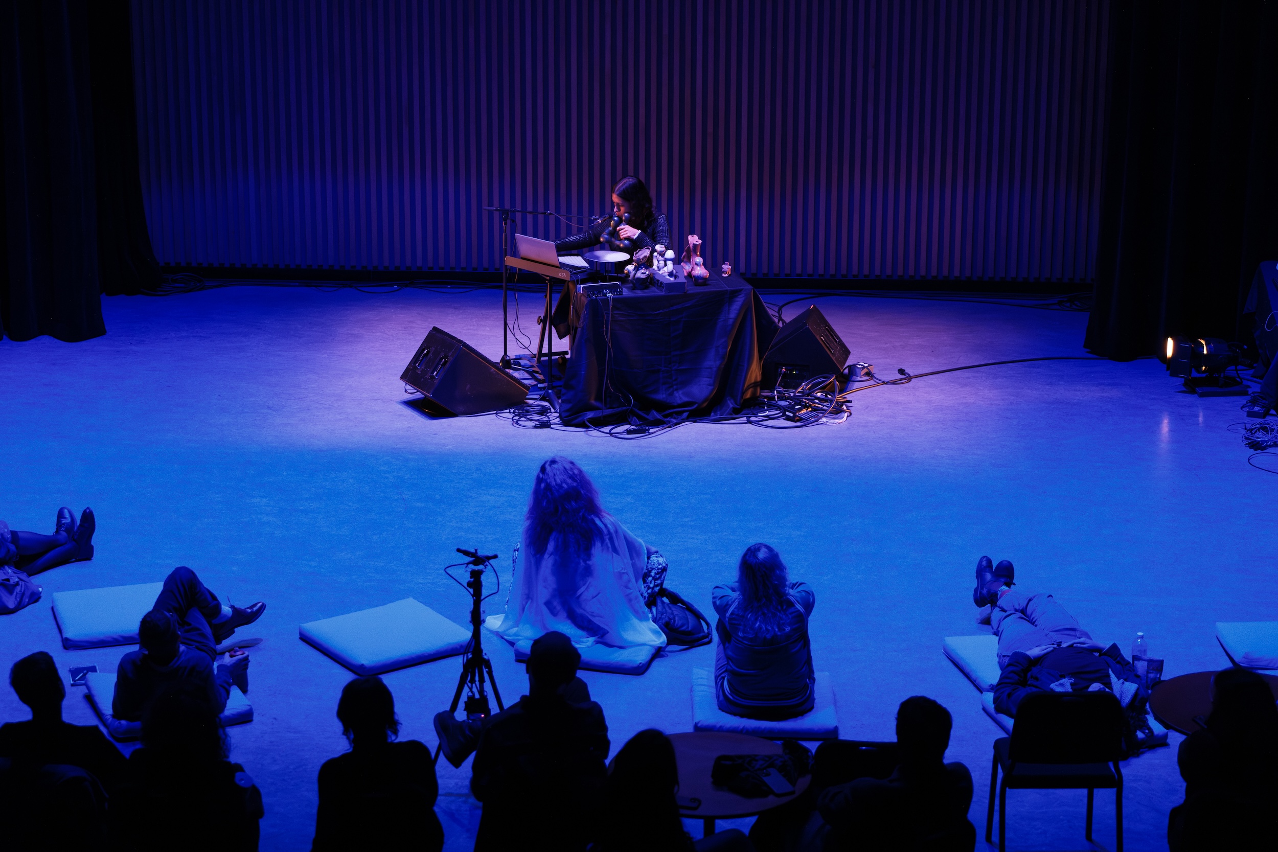 Vica Pacheco sits at a table with electronic equipment on stage under blue lighting while an audience watches, some seated on chairs and others lying on cushions.