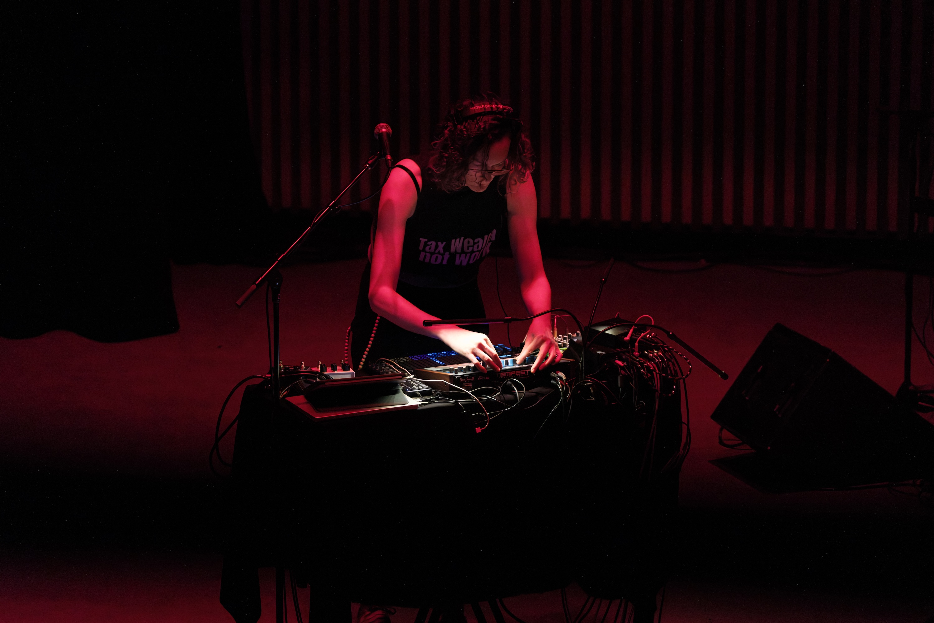 Brady Marks wearing headphones and a "Tax Wealth, Not Work" tank top playing music with a synthesizer on a dimly lit stage with red lighting.