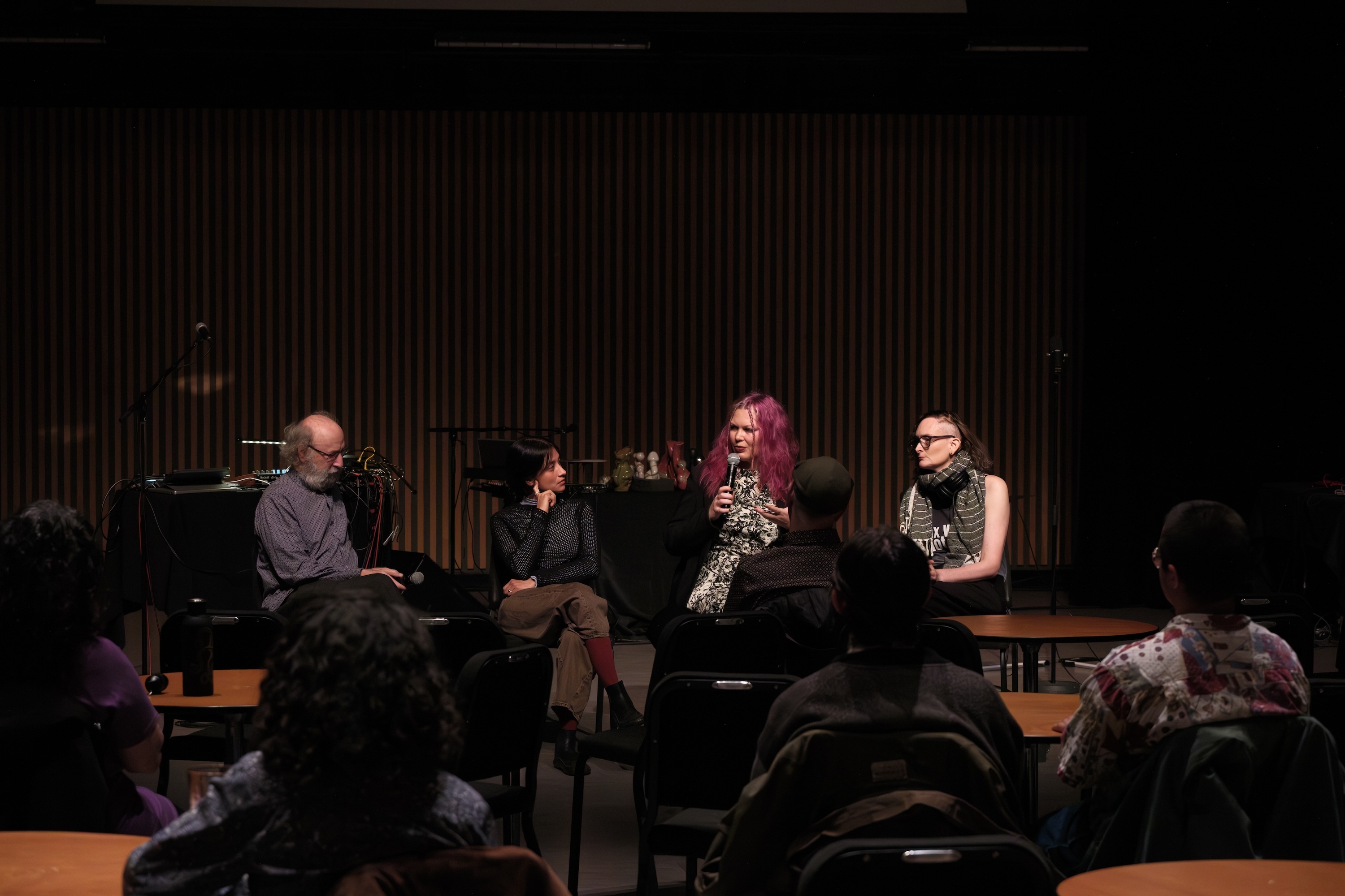 Brady Marks, Erin Gee, Vica Pacheco, and Giorgio Magnanensi sit on a panel in front of an audience, with Erin speaking into a microphone; tables and chairs are visible in the foreground.
