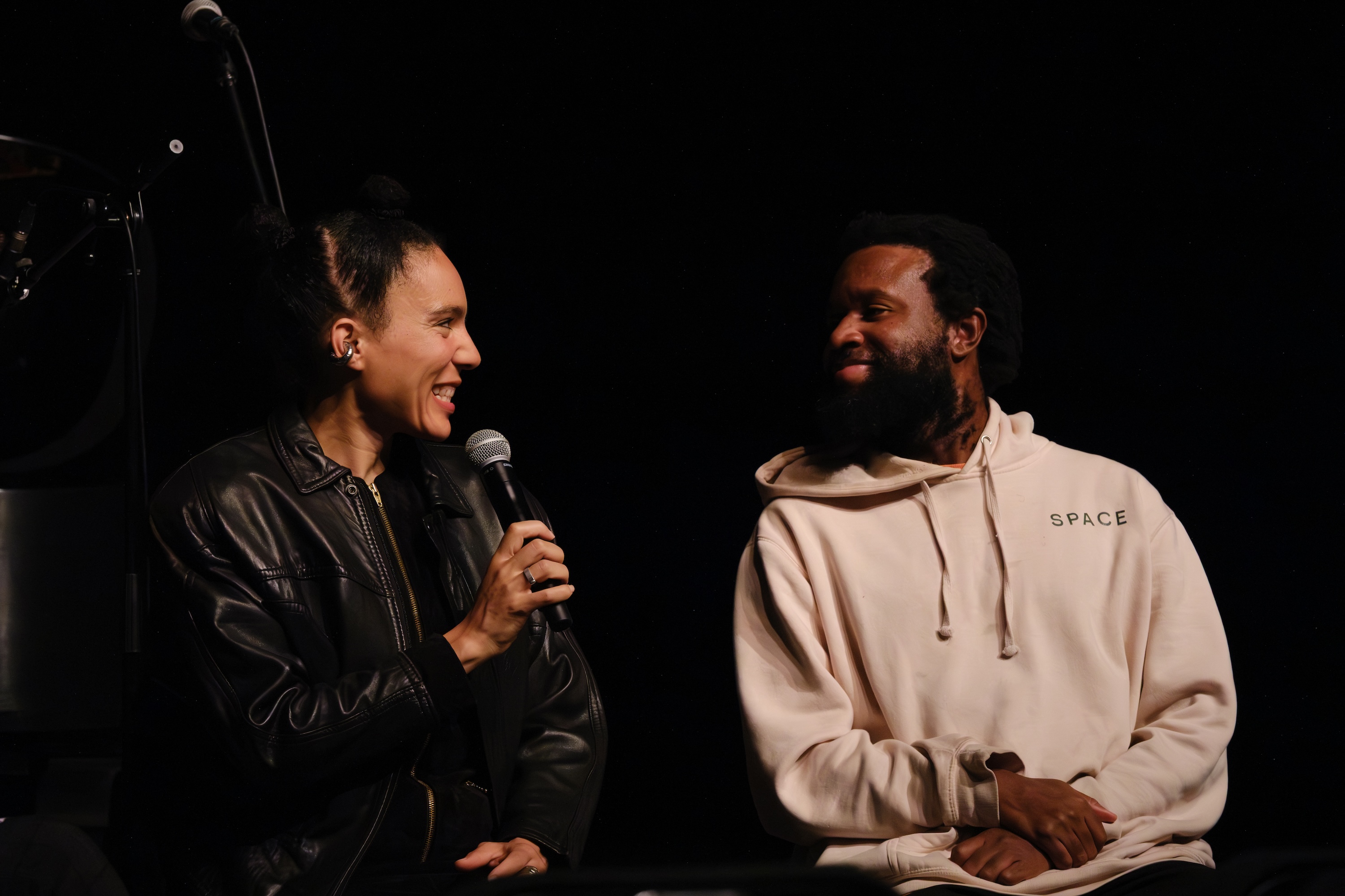 Anaïs Maviel holding a microphone speaks while smiling at JJJJJerome Ellis in a cream hoodie labeled "SPACE" against a dark background.