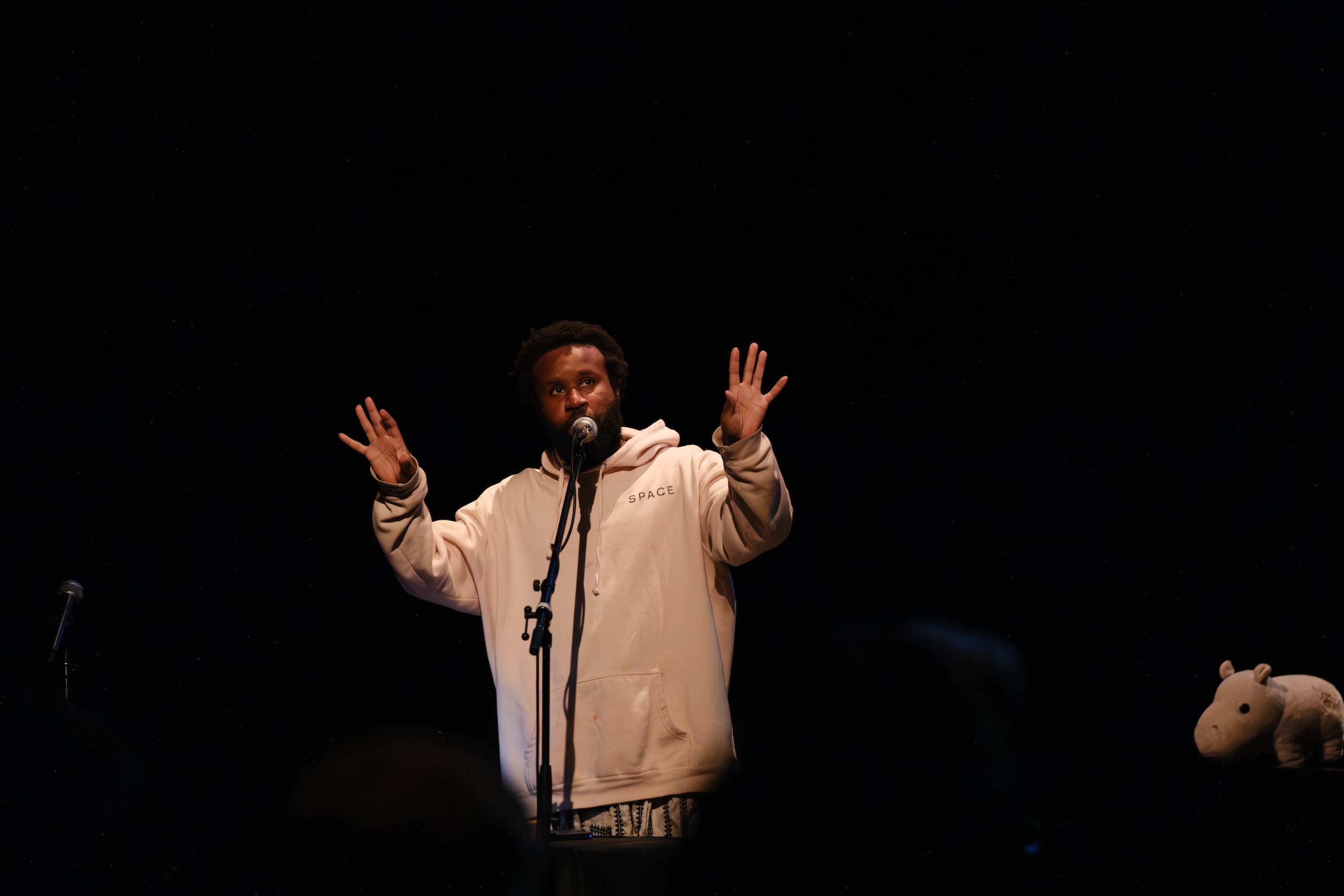 JJJJJerome Ellis in a light-colored hoodie speaks into a microphone on stage with raised hands; a stuffed hippo toy sits on a piano to the right.