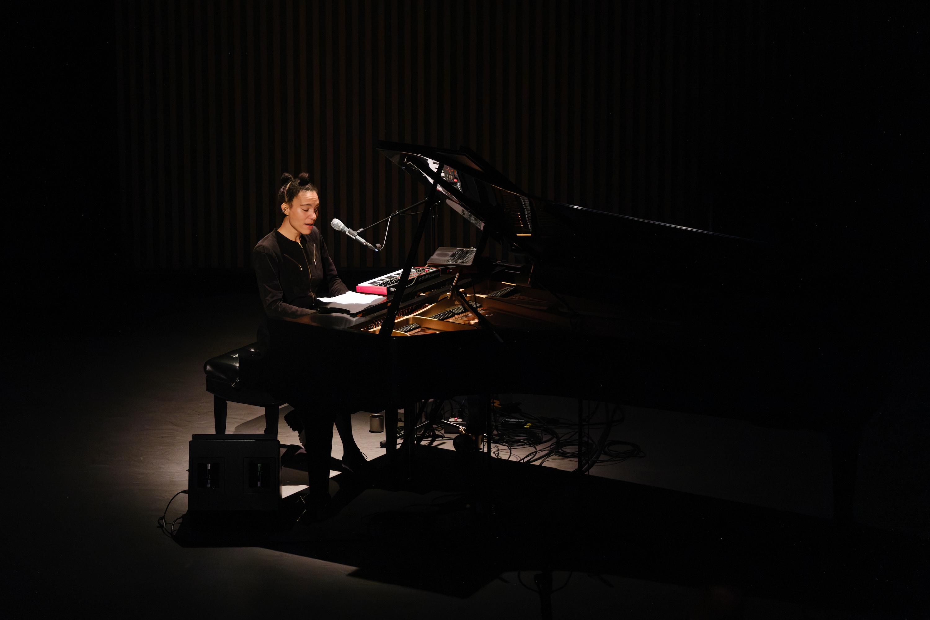 Anaïs Maviel sits at a grand piano on a dimly lit stage, playing and singing into a microphone.