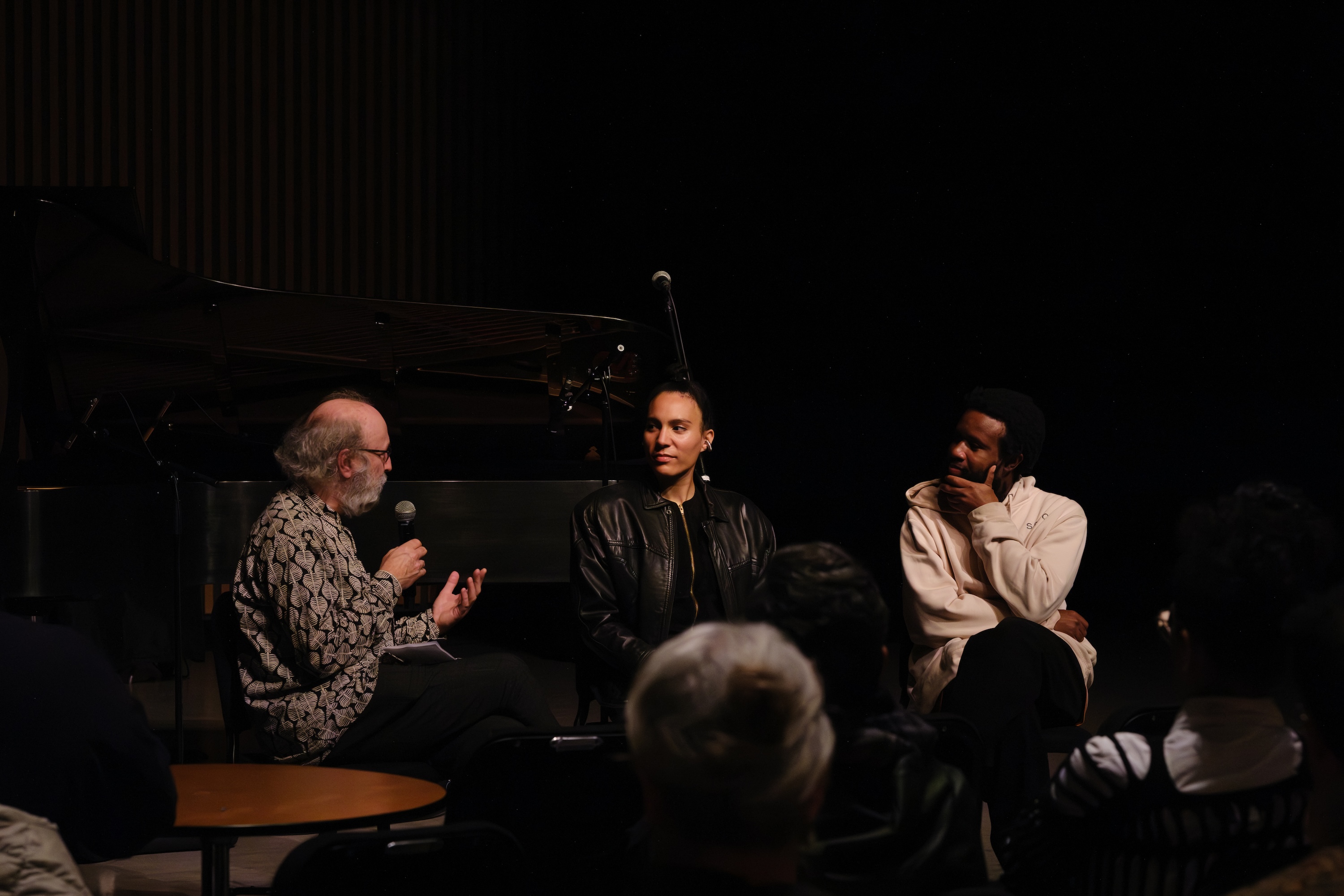 Anaïs Maviel, JJJJJerome Ellis, and Giorgio Magnanensi sit on stage in discussion; Giorgio holds a microphone, while Anaïs and JJJJJerome listen. A grand piano and microphone stand are in the background. Audience members are visible in front.