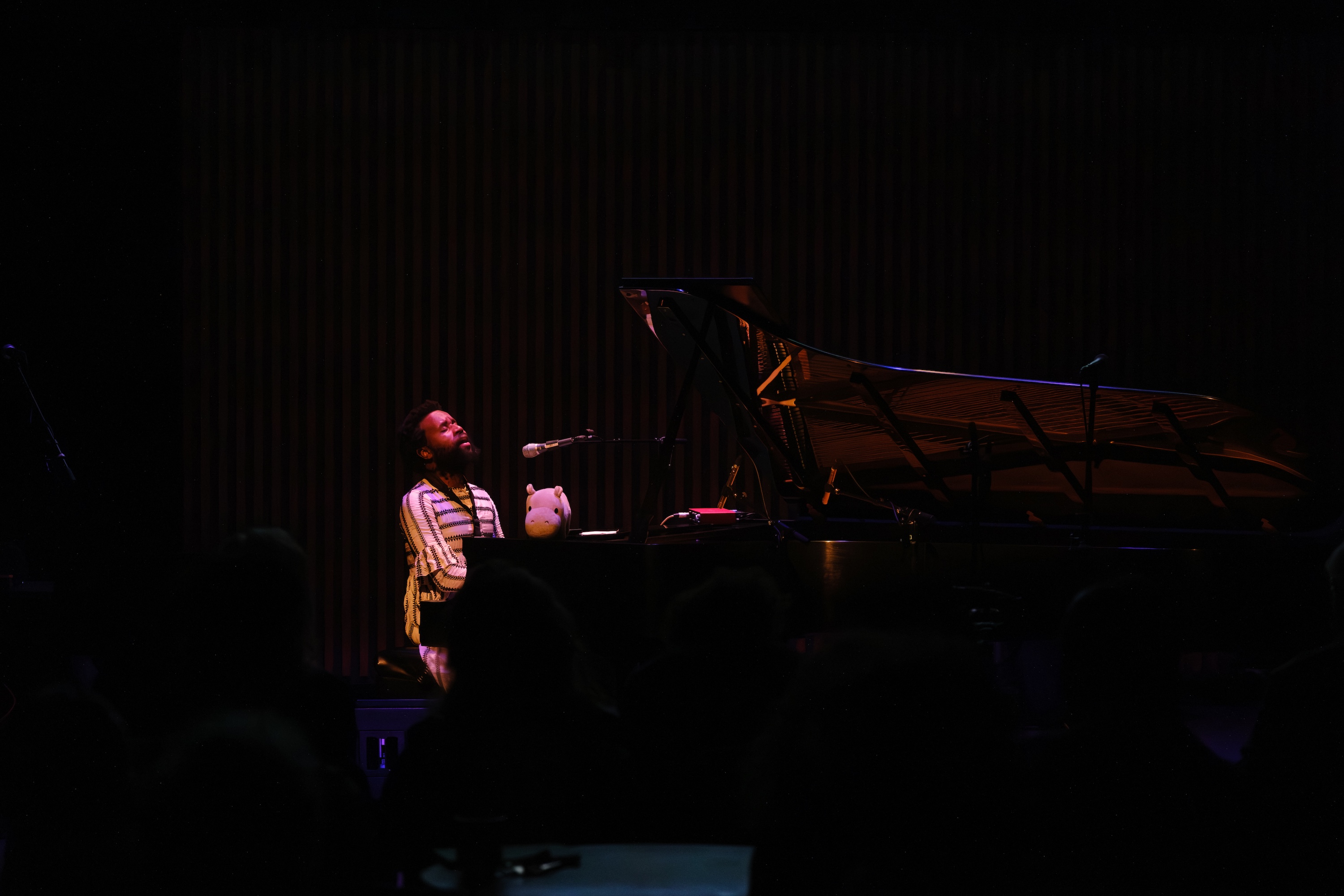 JJJJJerome Ellis sits at a grand piano on a dimly lit stage, singing into a microphone with an audience in the foreground. A stuffed hippo toy sits on the piano.