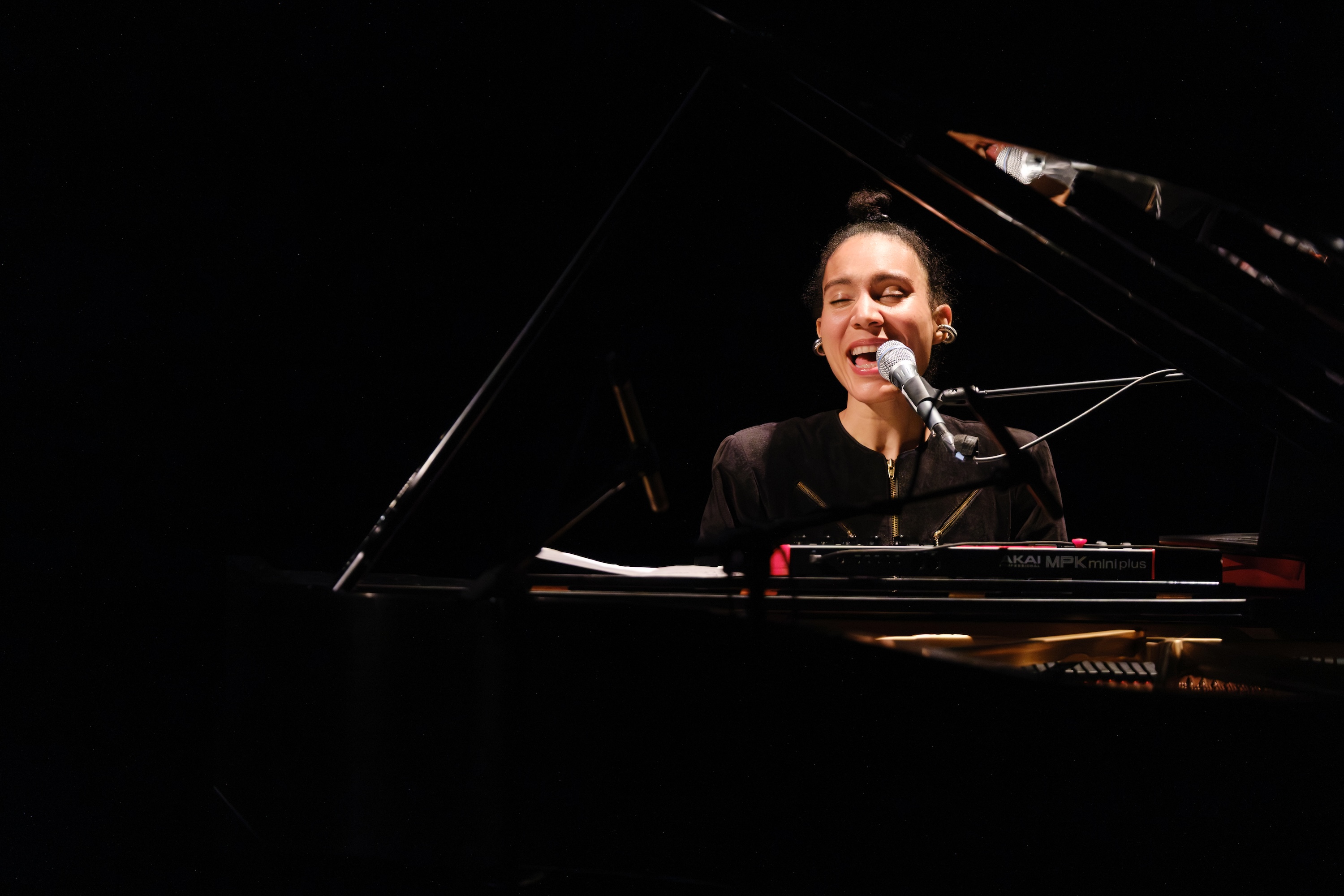 Anaïs Maviel sits at a grand piano, singing into a microphone on stage, illuminated by stage lighting against a dark background.