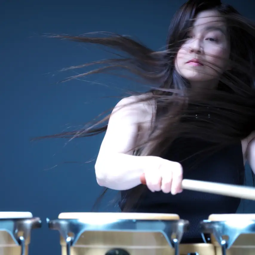 Kuniko Kato, a Japanese percussionist, stands to the right behind a set of drums. She wears a black tank top, and her long brown hair flows in the wind.
