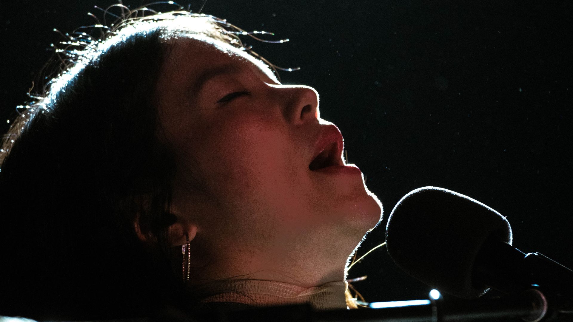 Close-up profile of a singer with closed eyes and an open mouth, illuminated against a dark background, microphone visible.