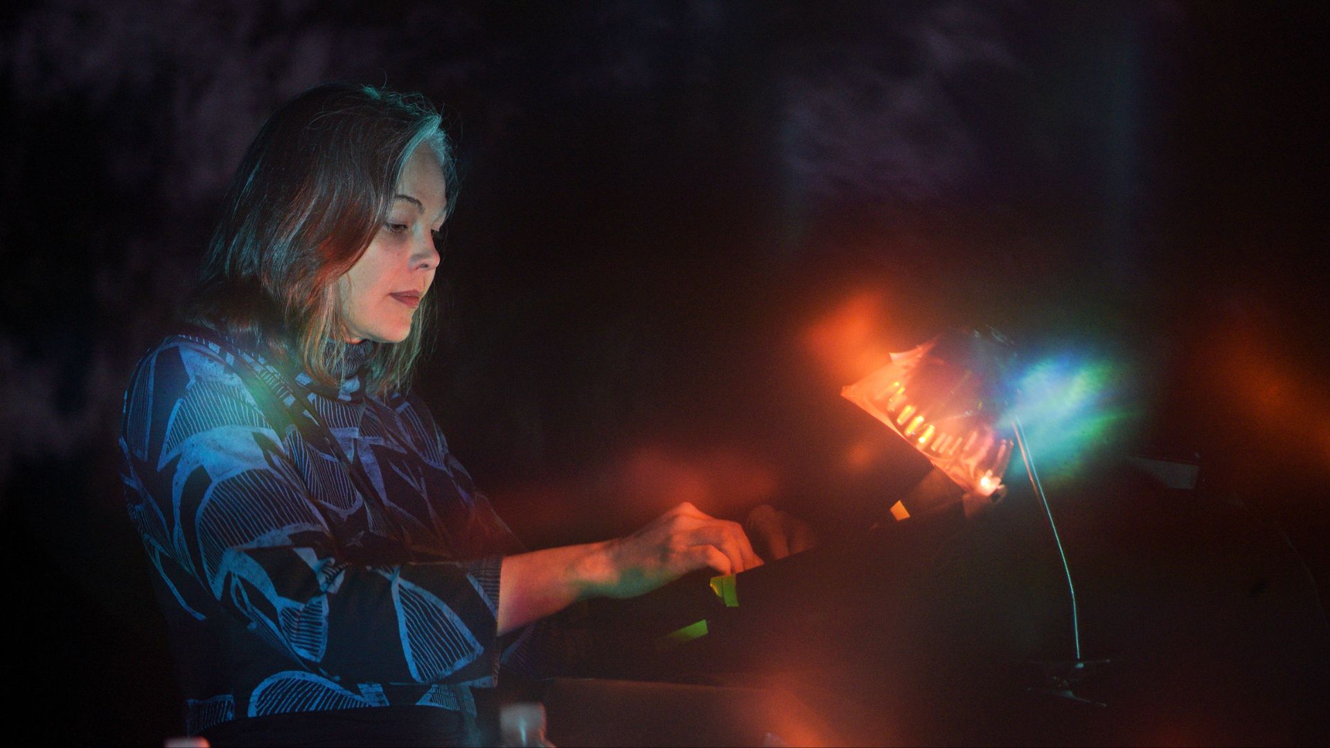 A woman with medium-length brown hair plays an electronic instrument, illuminated by colorful stage lights in a dim setting. She wears a dark top with a blue abstract pattern.