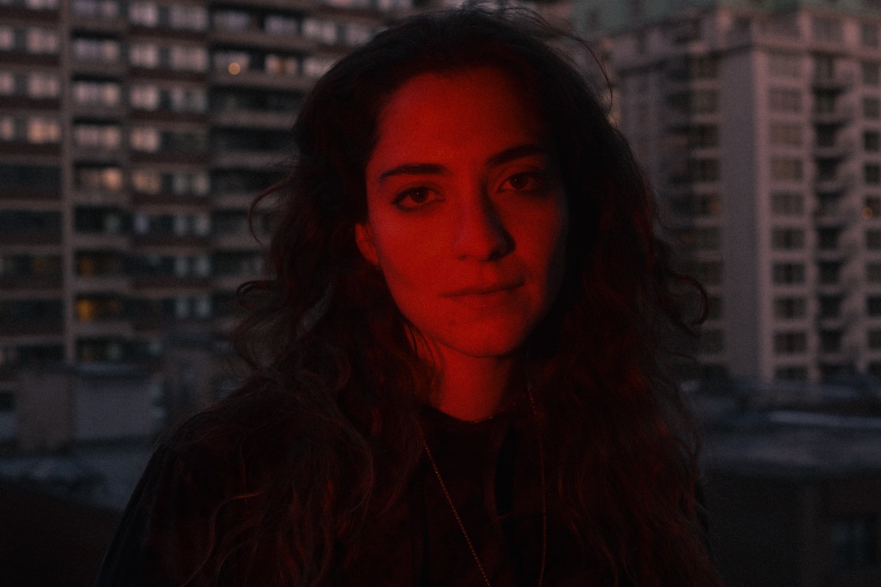 Close-up portrait of a woman (Nadah El Shazly) with dark wavy hair illuminated by red light, set against a blurred urban background.