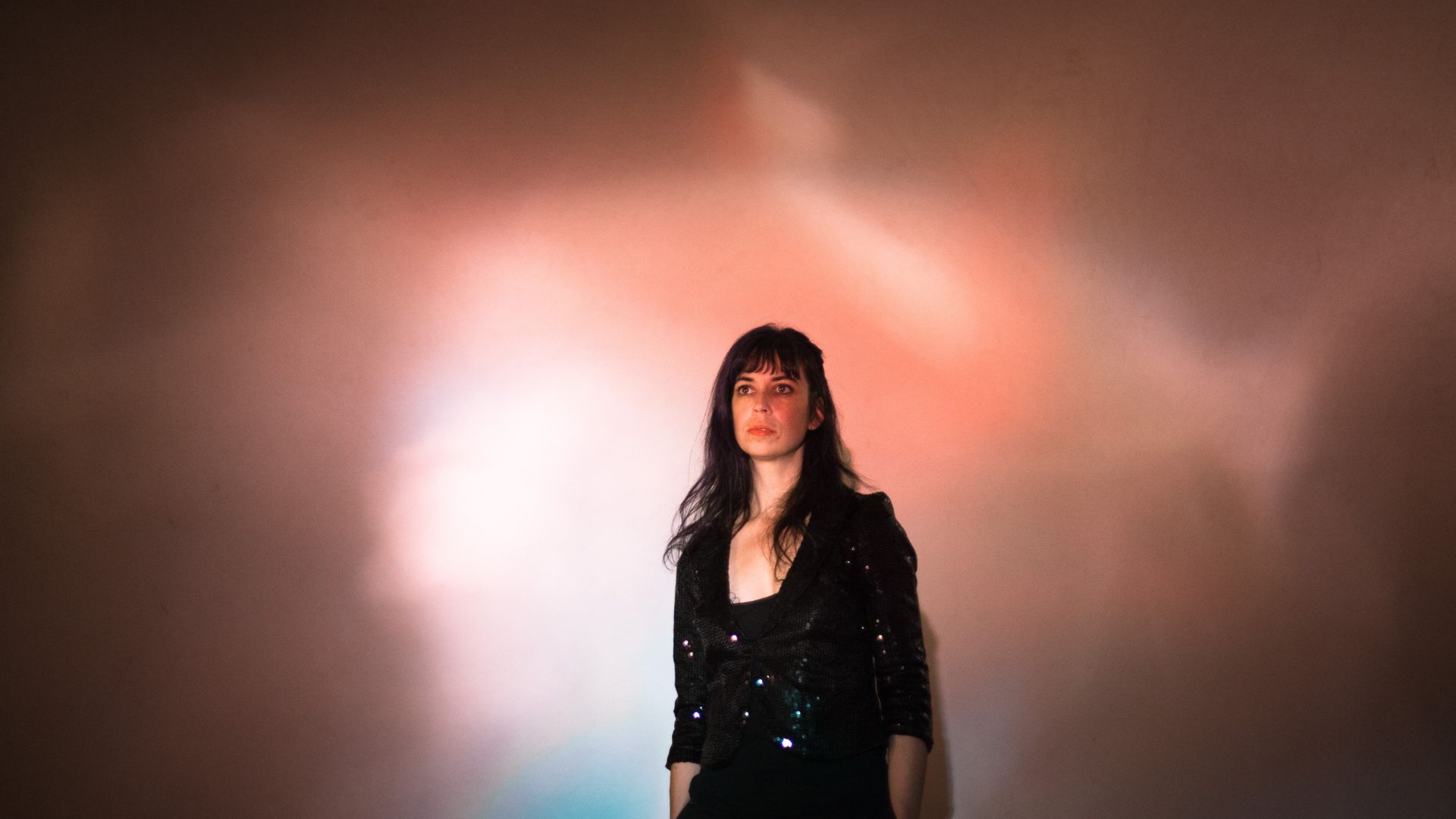 A woman with dark hair stands in front of a softly lit, colorful background, wearing a black sequined jacket. Her expression is serious as she looks directly at the viewer.