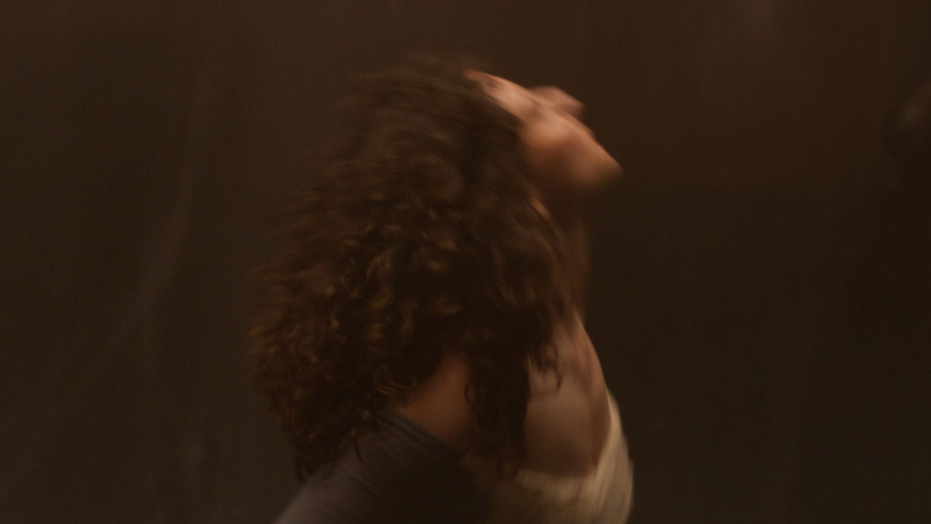 A dynamic still of a woman with curly hair moving energetically against a warm brown background, her head tilted back and eyes closed, conveying emotion.