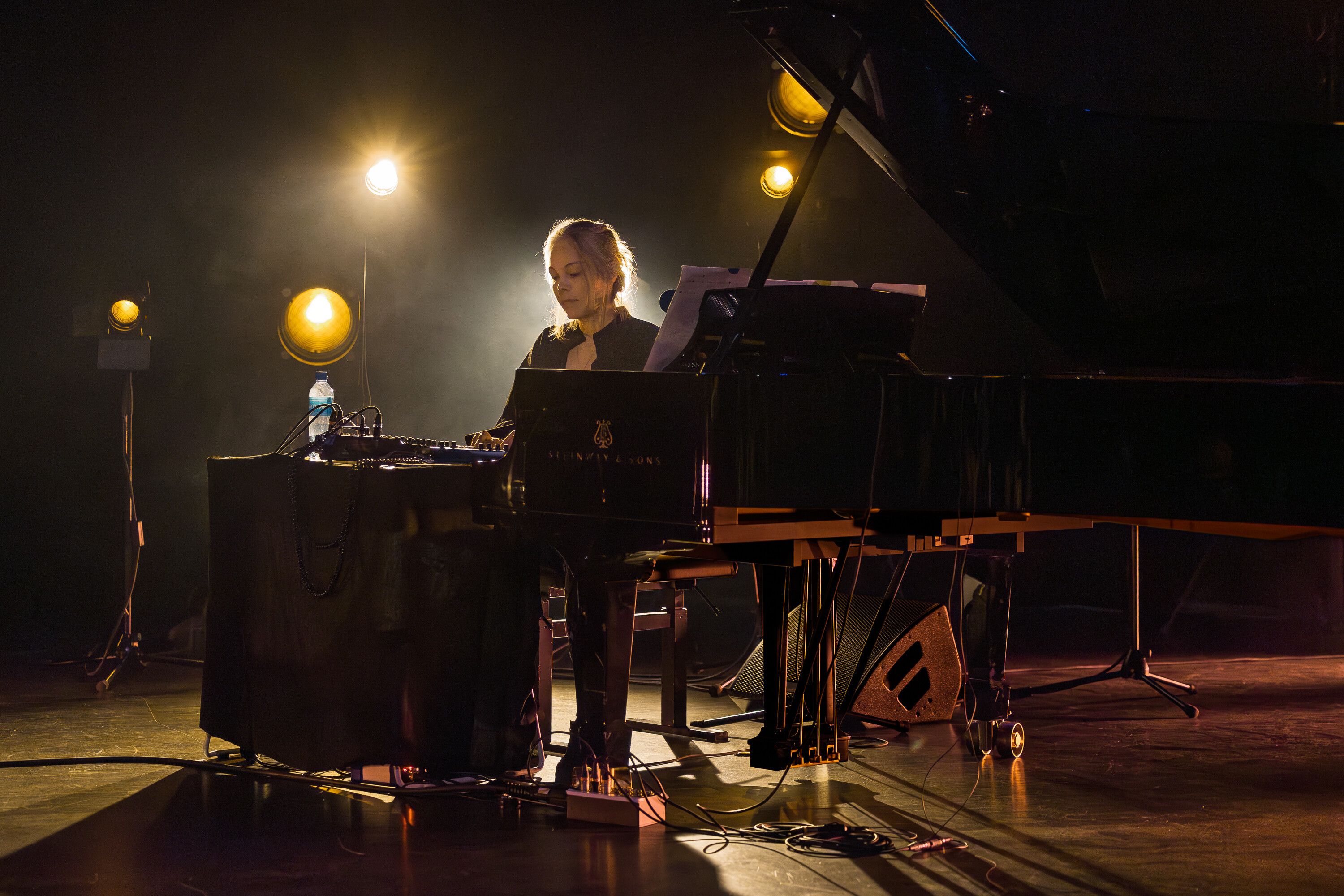 A woman, Olivia Block, plays a grand piano on stage, surrounded by dramatic lighting and cables, creating a serene atmosphere during a performance.
