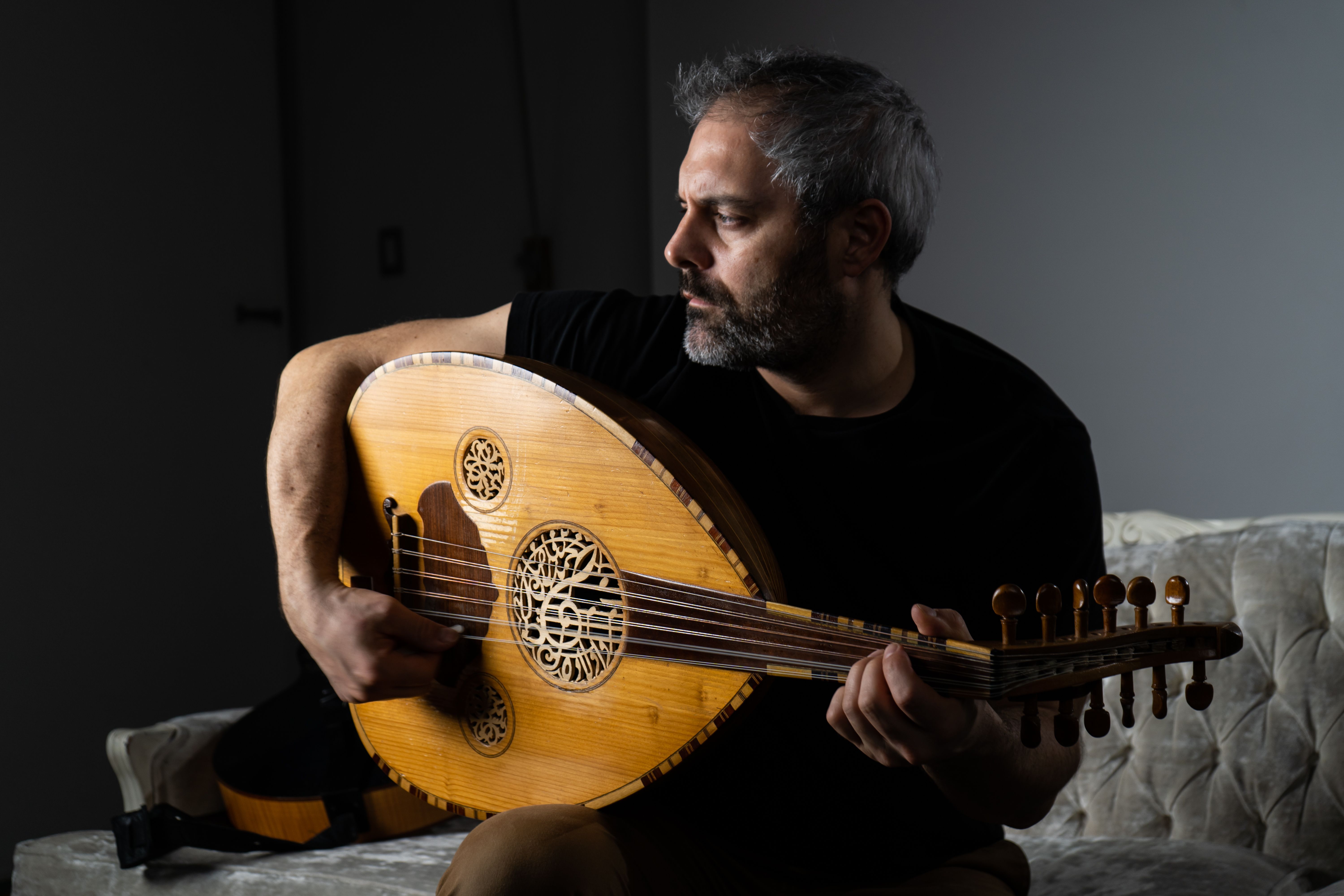 A bearded man in black plays a honey-colored oud while seated in a dimly lit room.