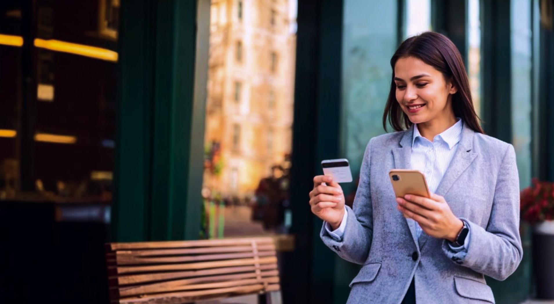 Smiling woman in a gray blazer holding a credit card and a smartphone outdoors.