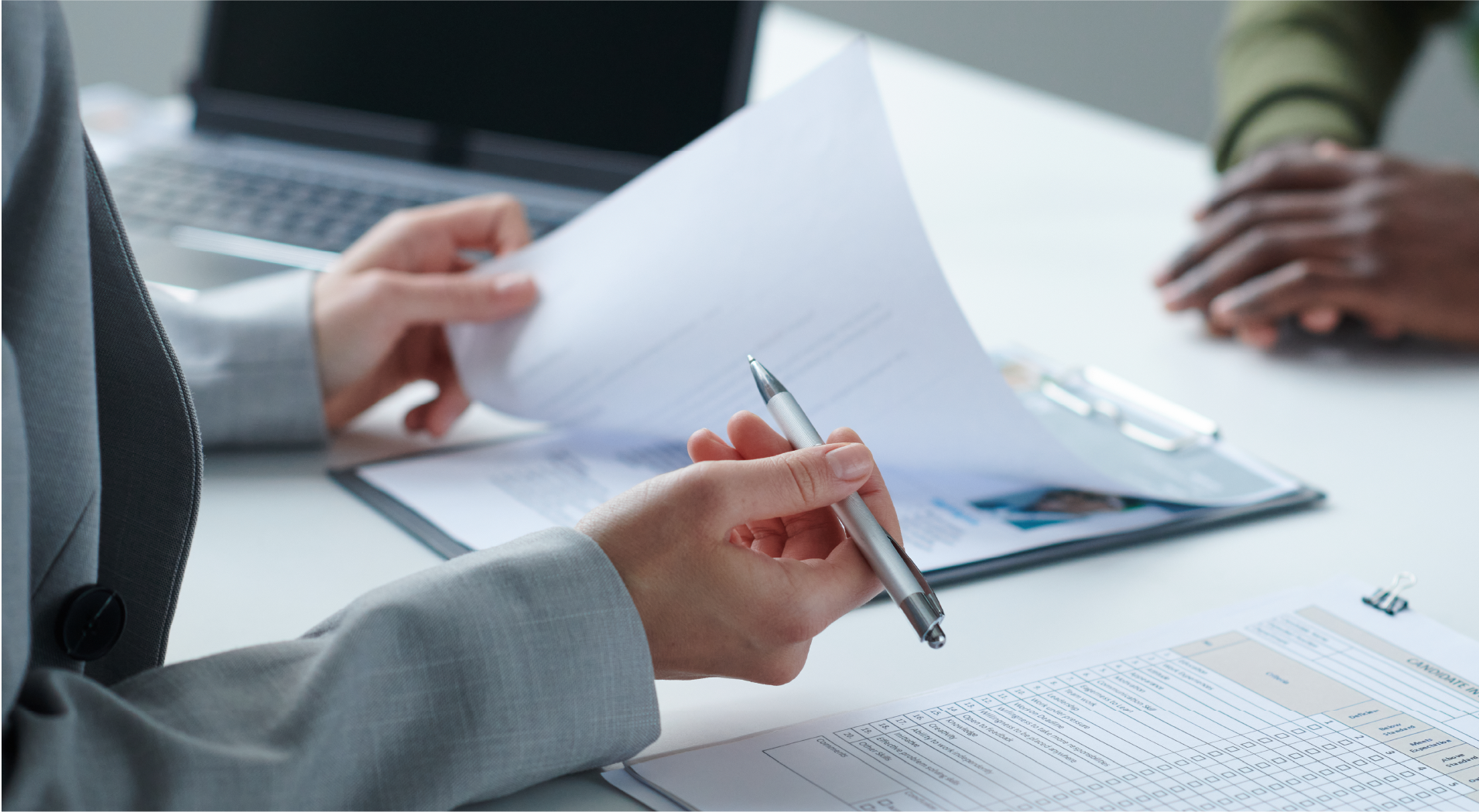 Person in a gray suit holding a pen and reviewing papers, including a clipboard with a candidate information form, during a meeting.