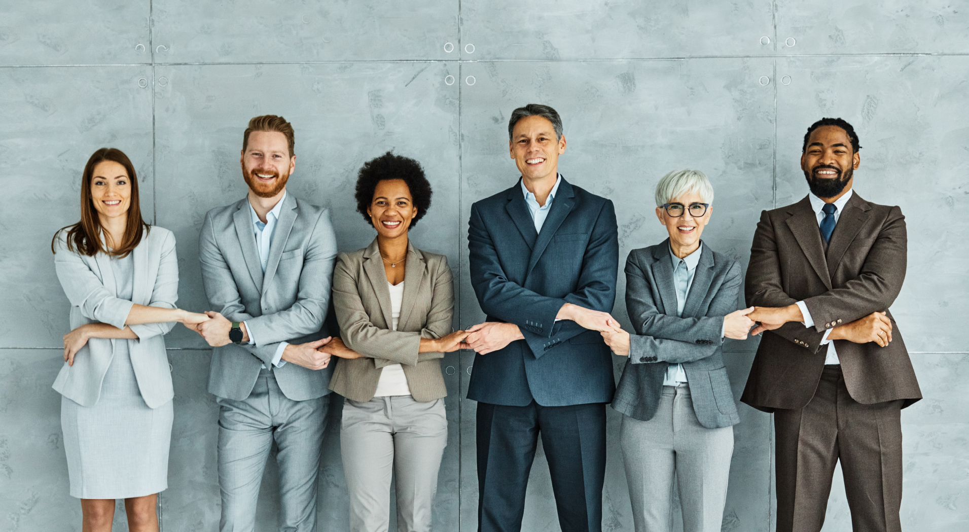Six diverse business professionals standing in a row smiling and linking arms in front of a gray wall.
