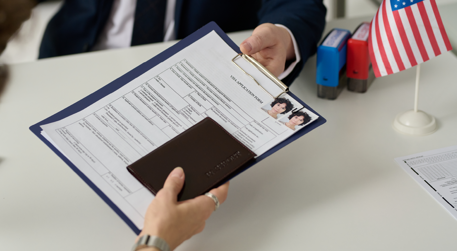 Person handing over a visa application form and passport cover to a man in a suit at a desk with a small American flag and stamps.