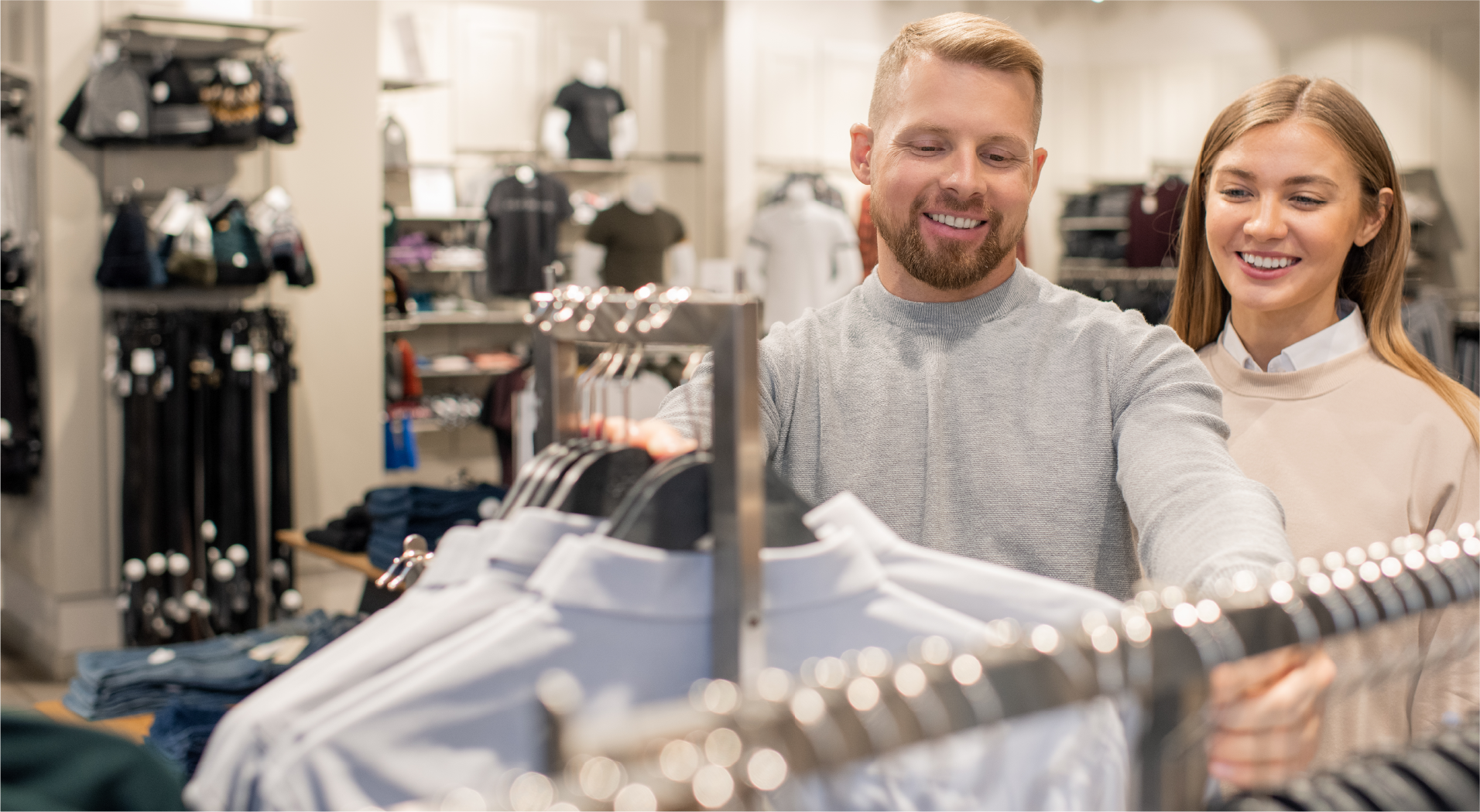 A smiling man and woman shopping for clothes, browsing shirts on a rack inside a retail store.